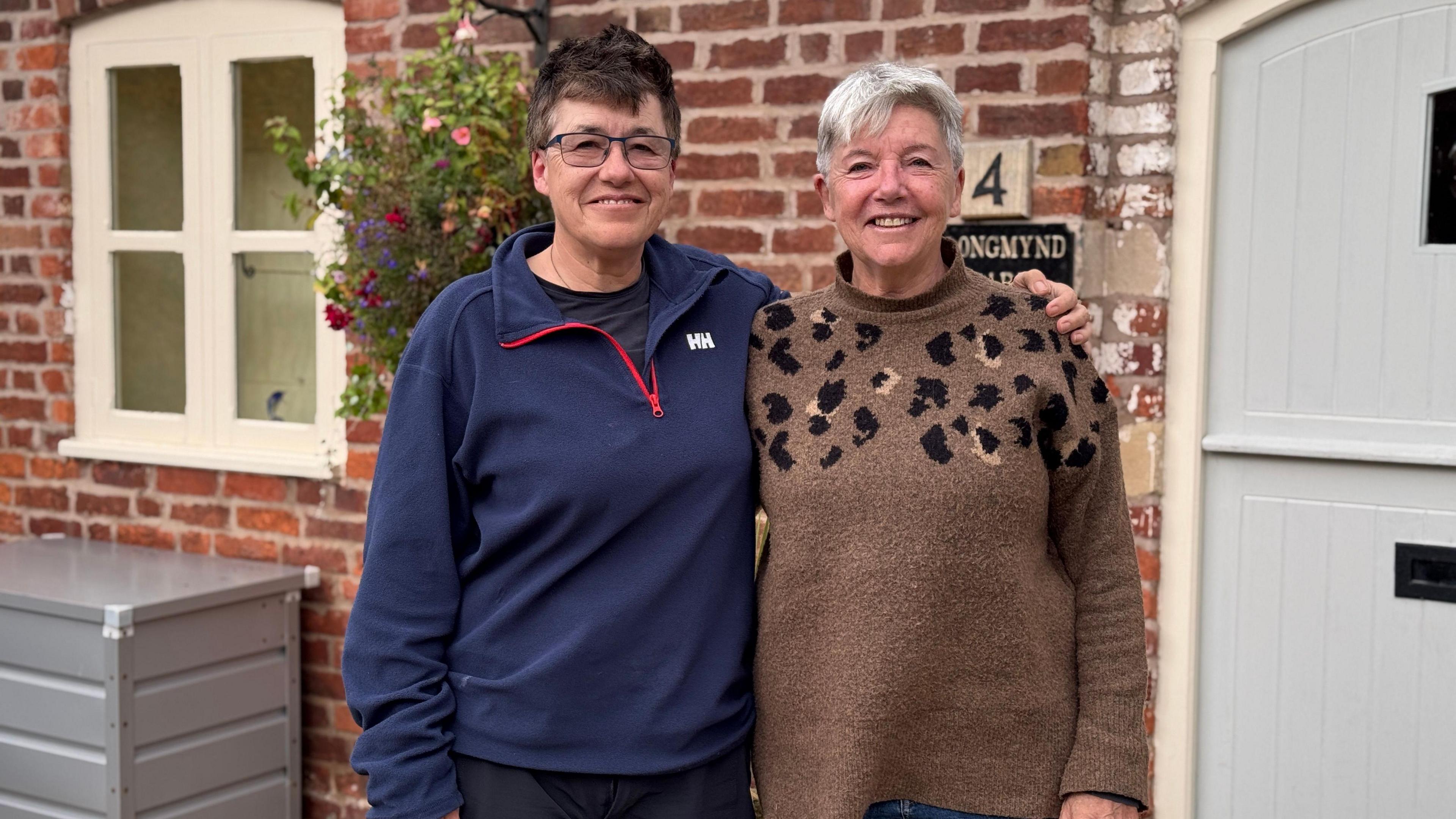 Two women are standing next to each other. The one on the left has short dark hair and is wearing glasses and a blue zipped jumper. The woman on the left has short grey hair and is wearing a brown-patterned jumper. They are standing in front of a brick-built house with a grey door. There is a large grey box and flowers nearby.