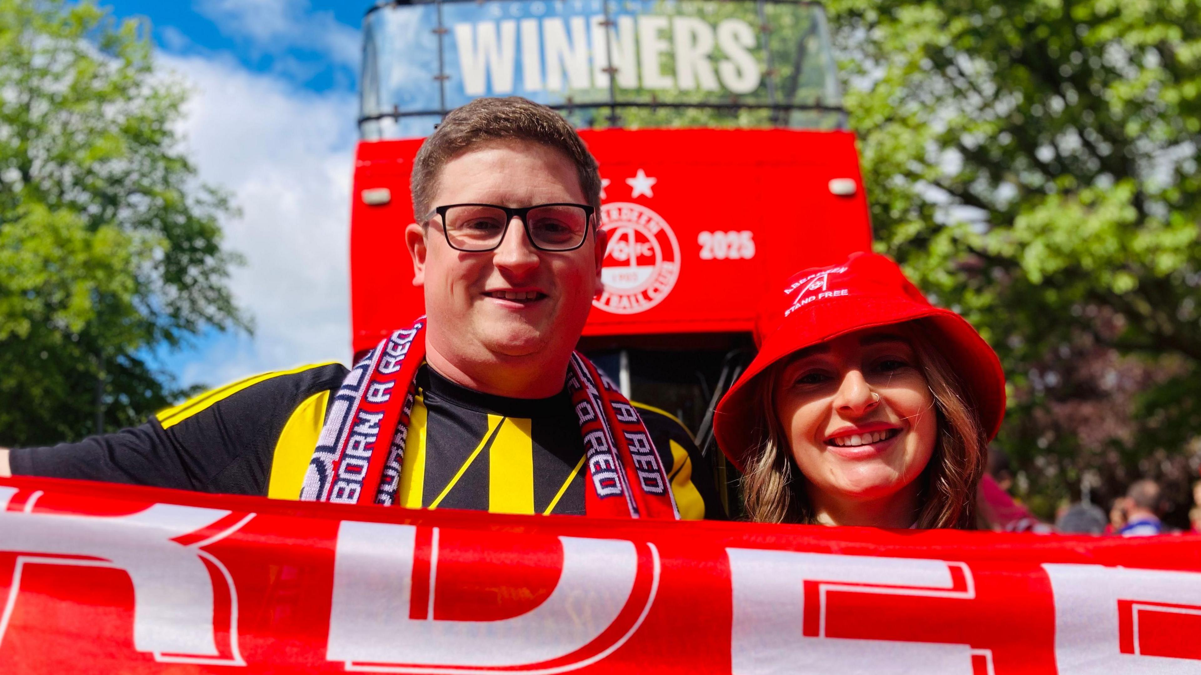 A man and woman in aberdeen hats, holding a scarf out in front of a bus.