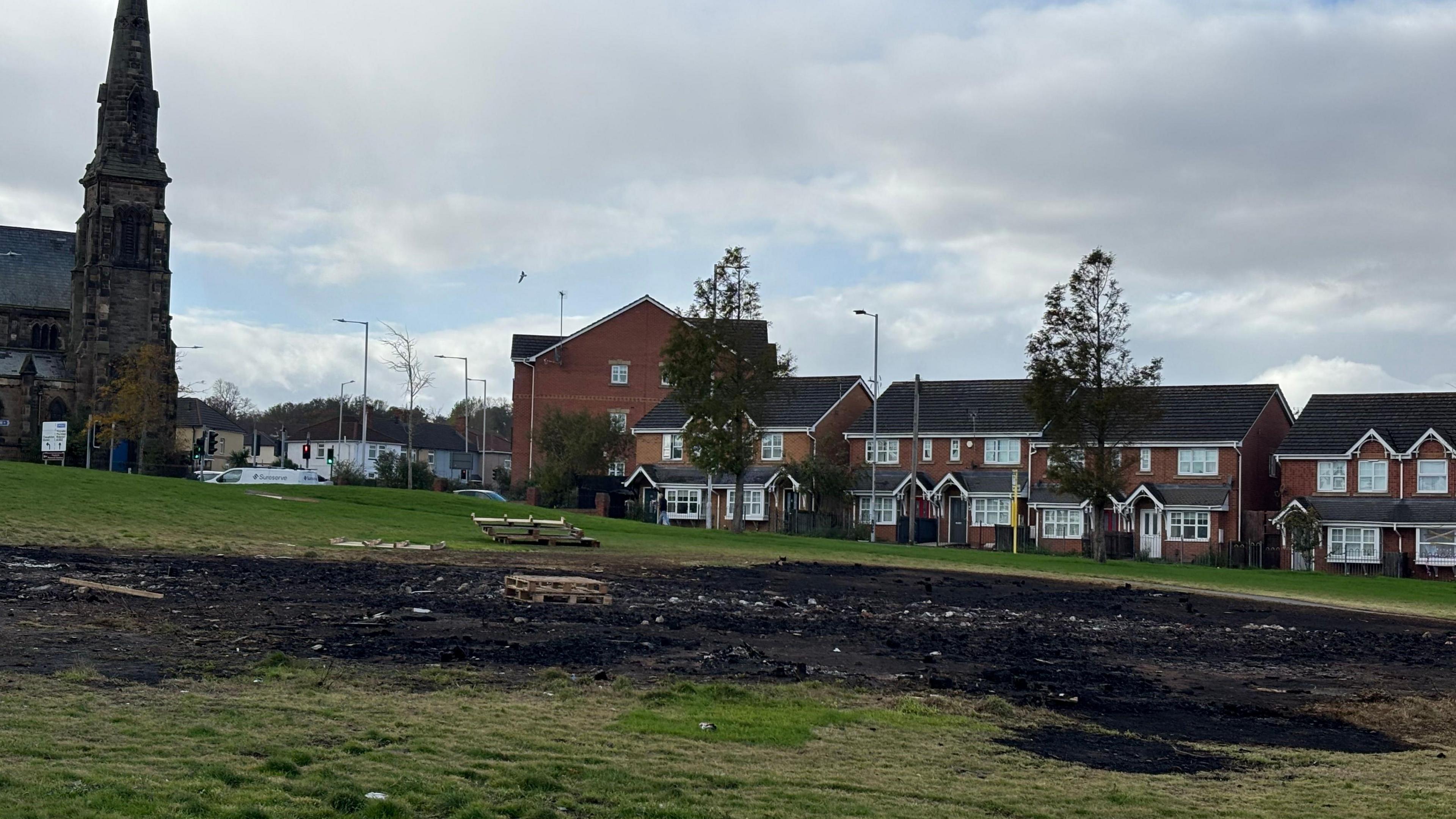 A large patch of charred ground in the middle of a field. A church can be seen in the background and houses line the road behind the field.