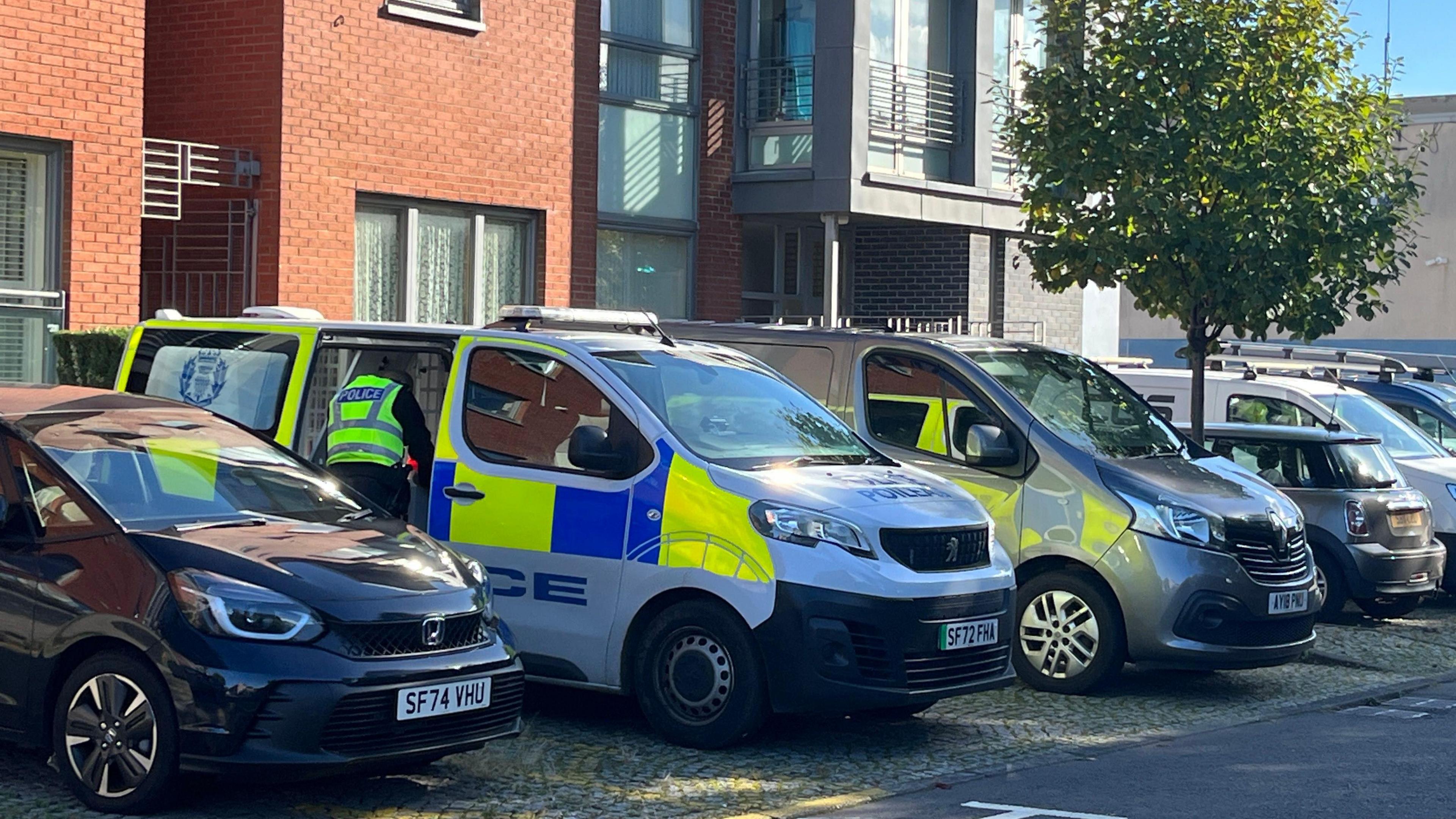 A police van parked outside a modern block of flats. An officer has the back door open and is either taking something out or placing something inside the vehicle. Several other cars and vans are parked nearby.