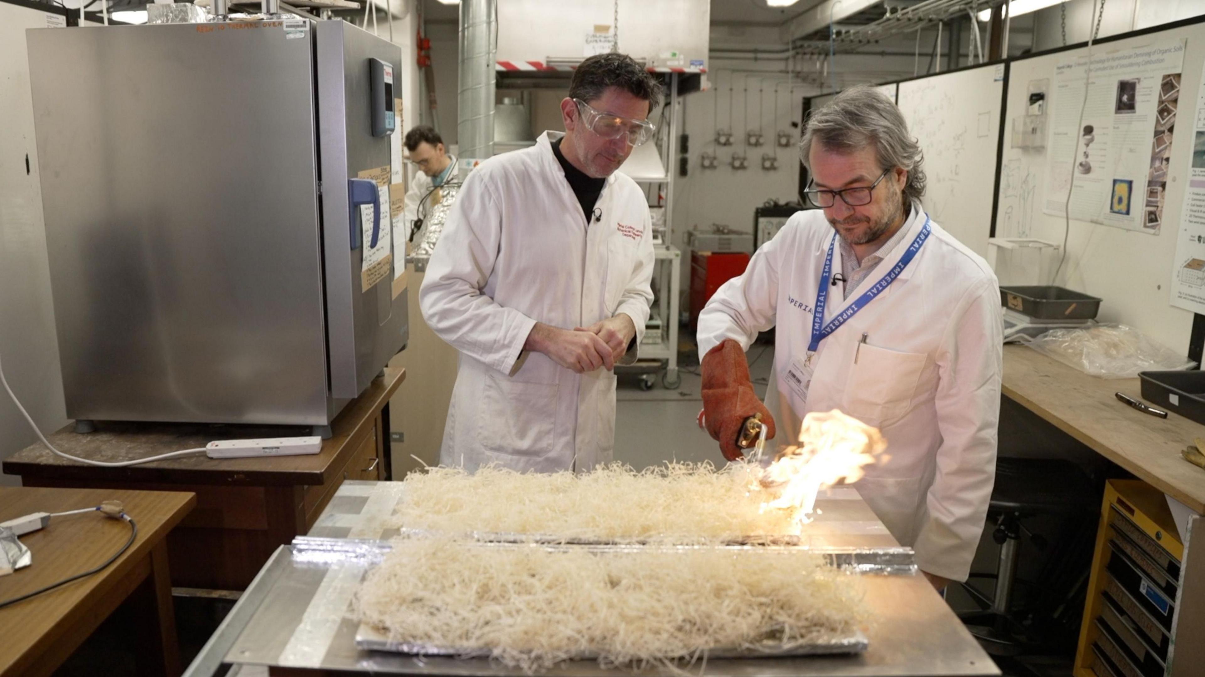 A professor with grey hair with a pair of glasses wearing a white overall coat is igniting dry straw on a steel plate with a fire burner in a laboratory. Justin Rowlatt - also wearing a white overall coat and a pair of safety glasses - is looking on next to the professor.  
