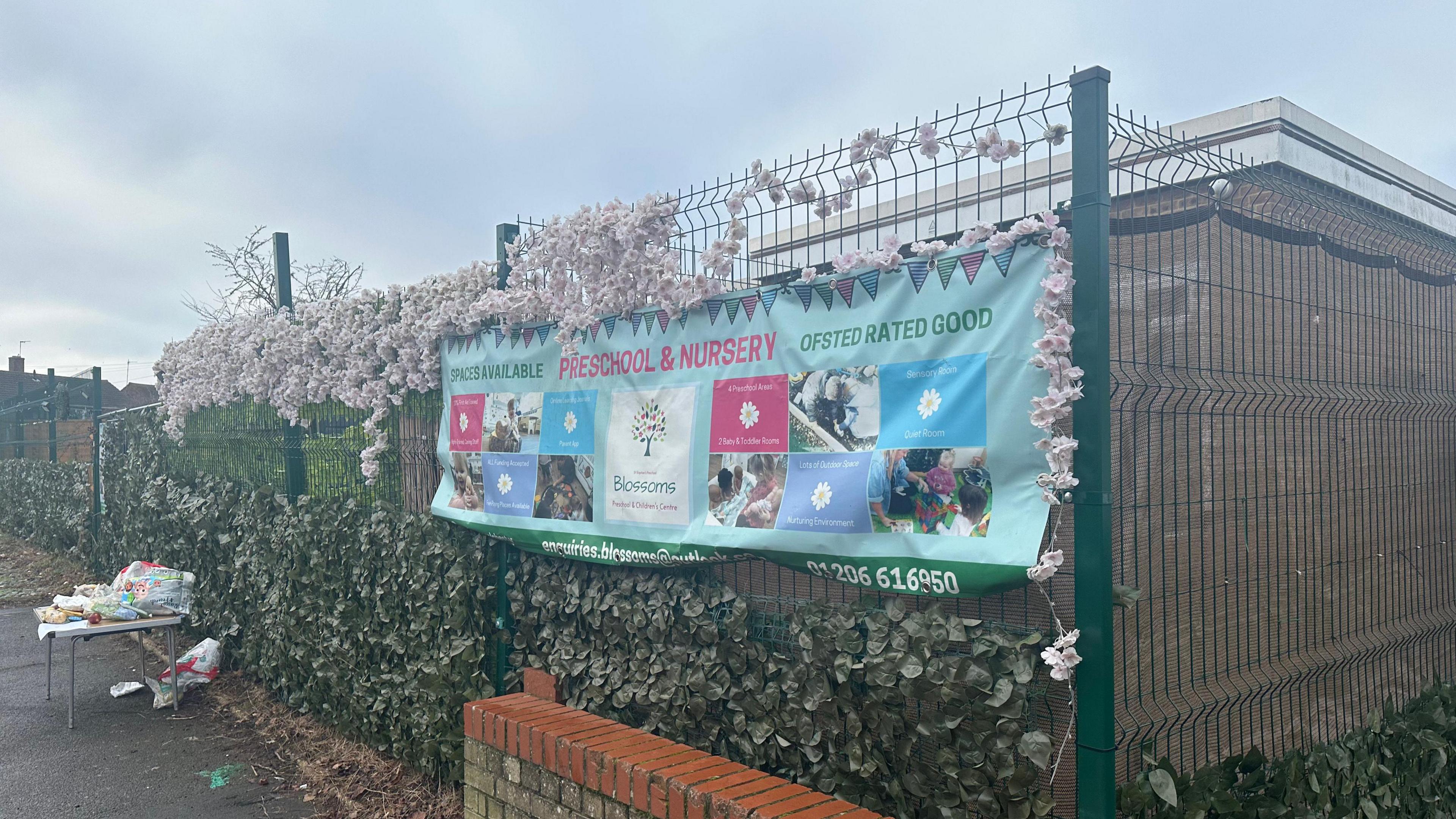 Another view of the side of Blossom Preschool and the banner that sits on its fence. A table sits outside the site with free food on offer.