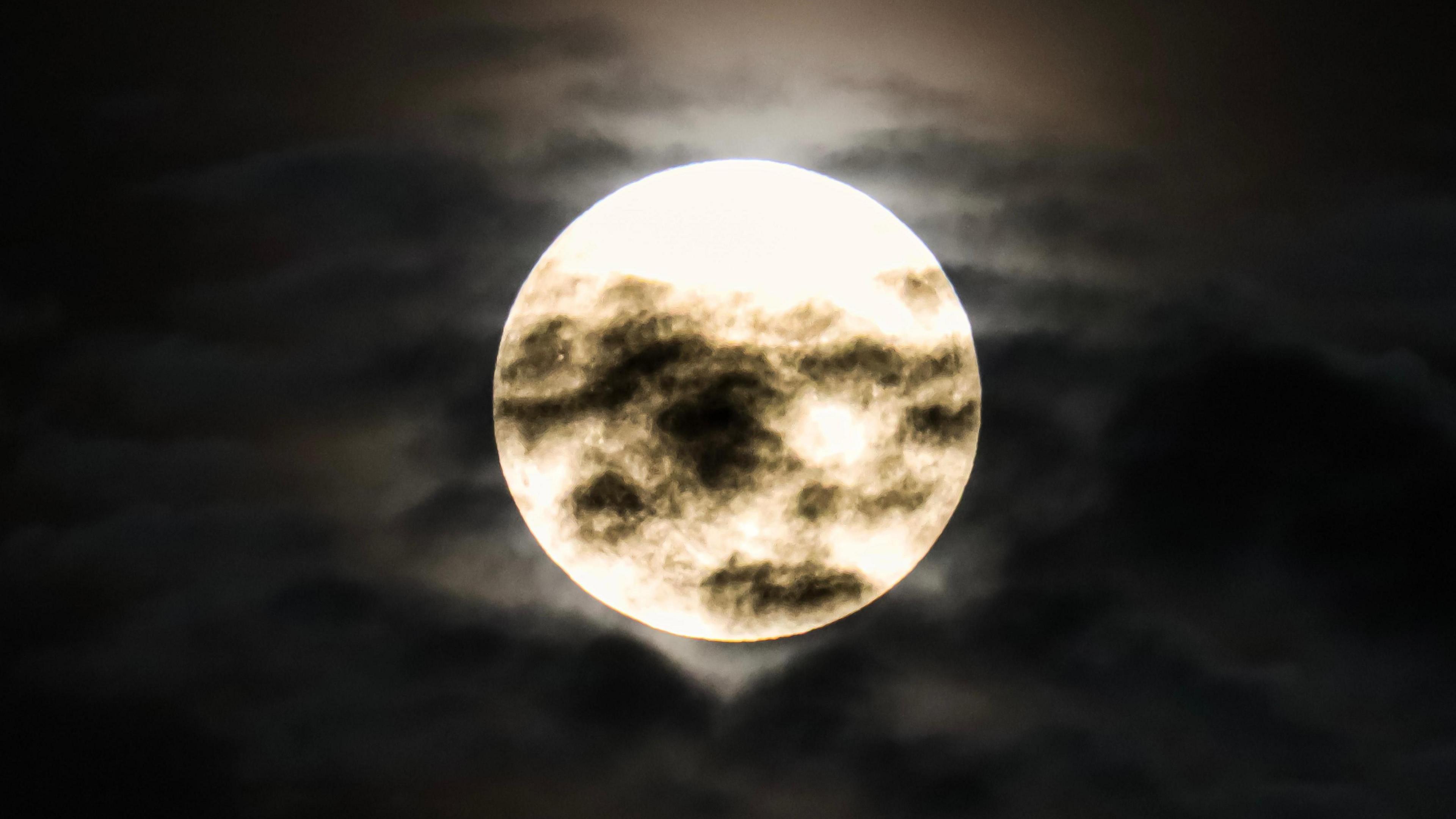 A large white Strawberry Moon in the night sky with black clouds crossing over it