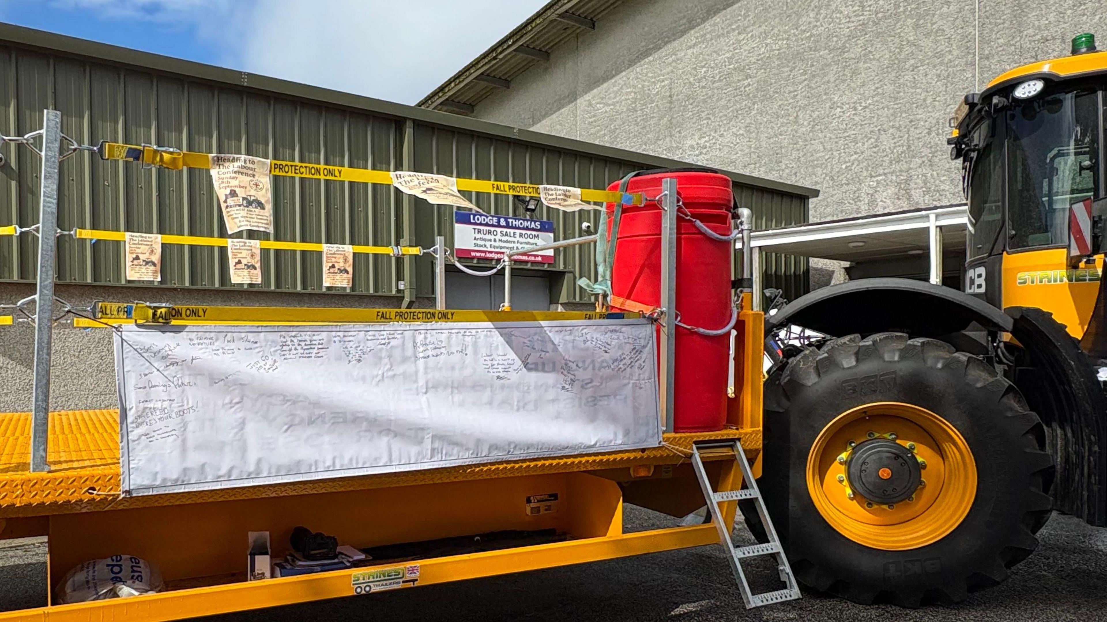 A banner with writing on it attached to a yellow trailer being towed by a yellow tractor