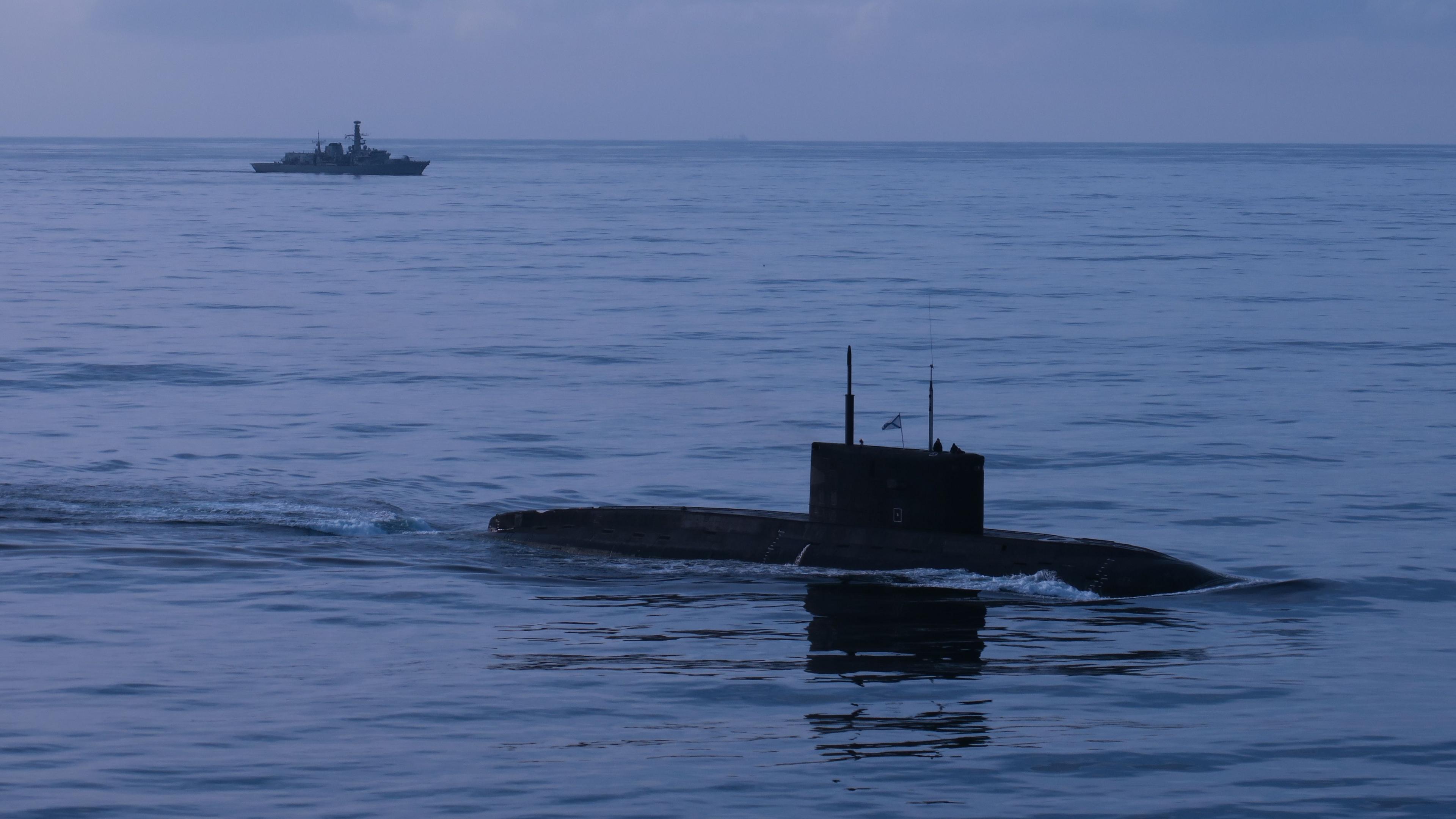 A submarine is seen in the water in the foreground, with a navy warship in the background