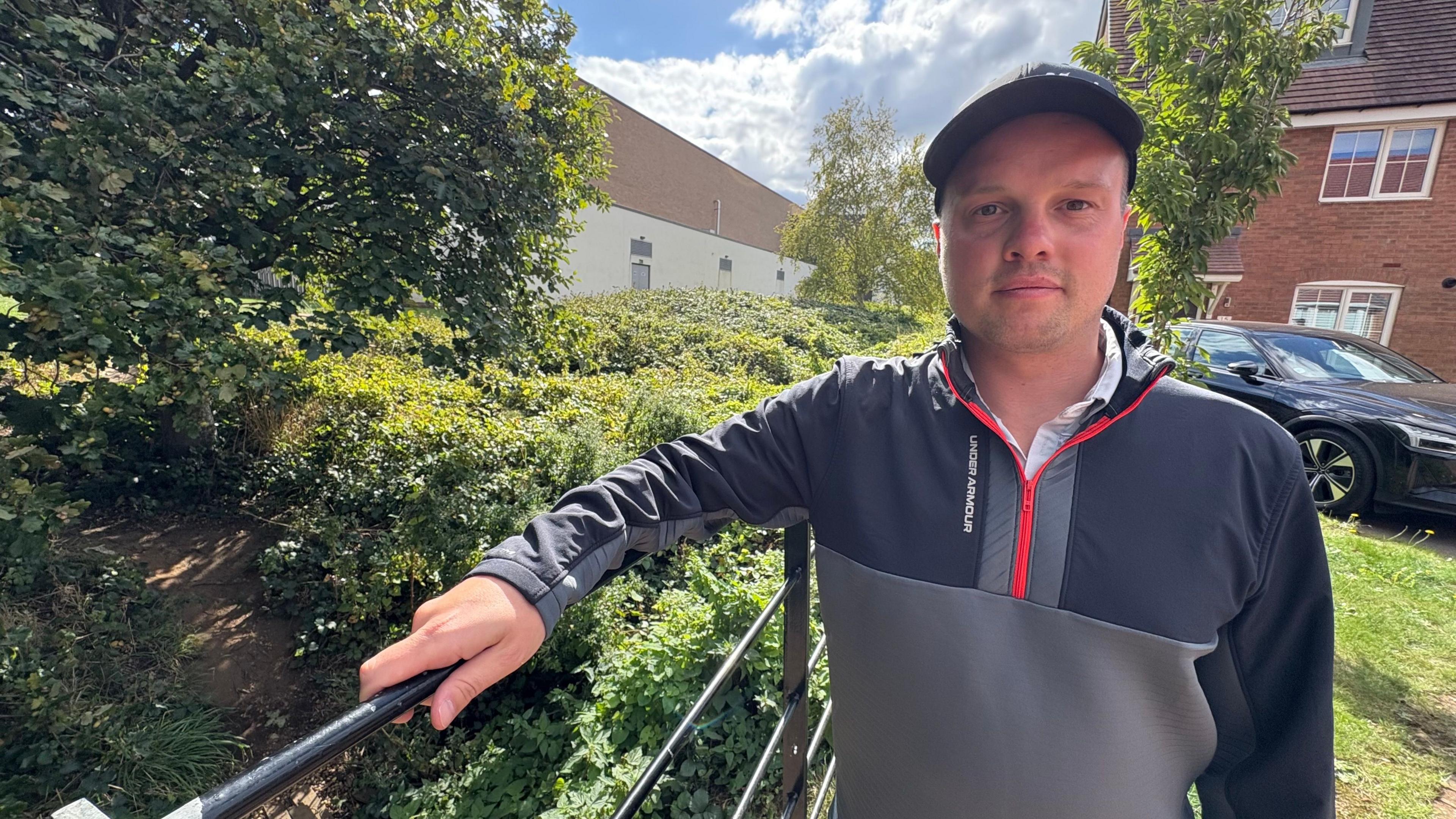 Connor Gladwin with dark-coloured cap and grey hoodie leaning on a black metal railing. There are green bushes behind him, and a leisure centre building is visible in the background. His own two-storey brick house and drive is visible to the right.