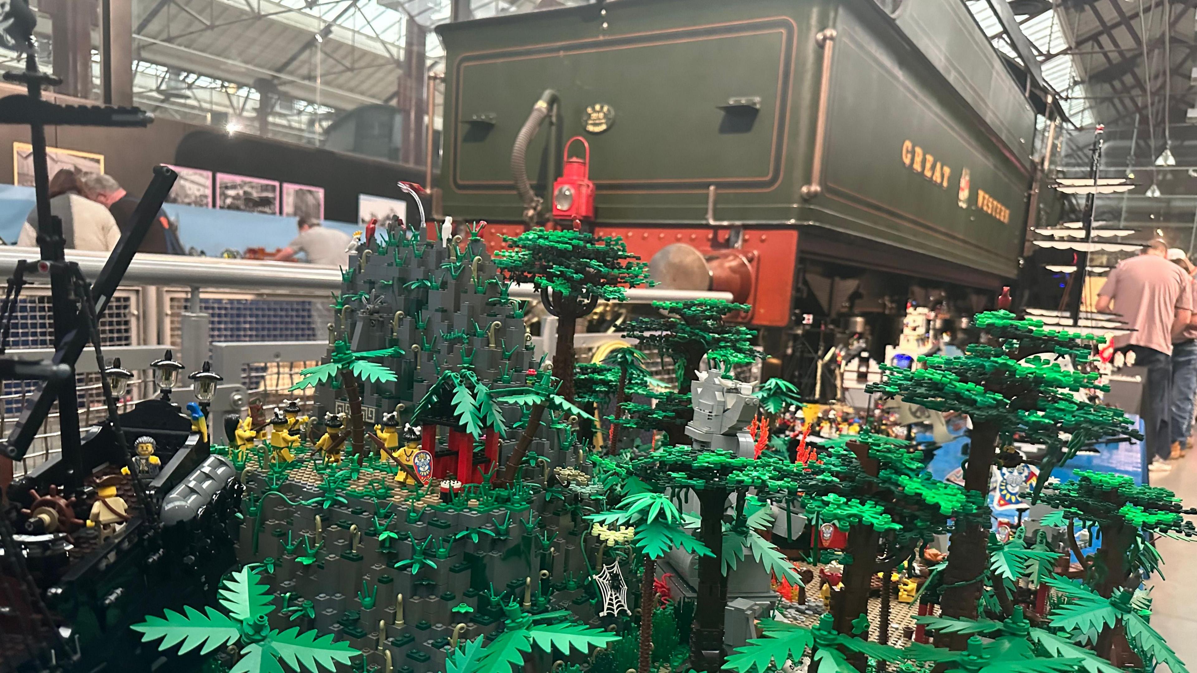 Lego trees are displayed in front of a historic locomotive in a museum. The locomotive is dark green.