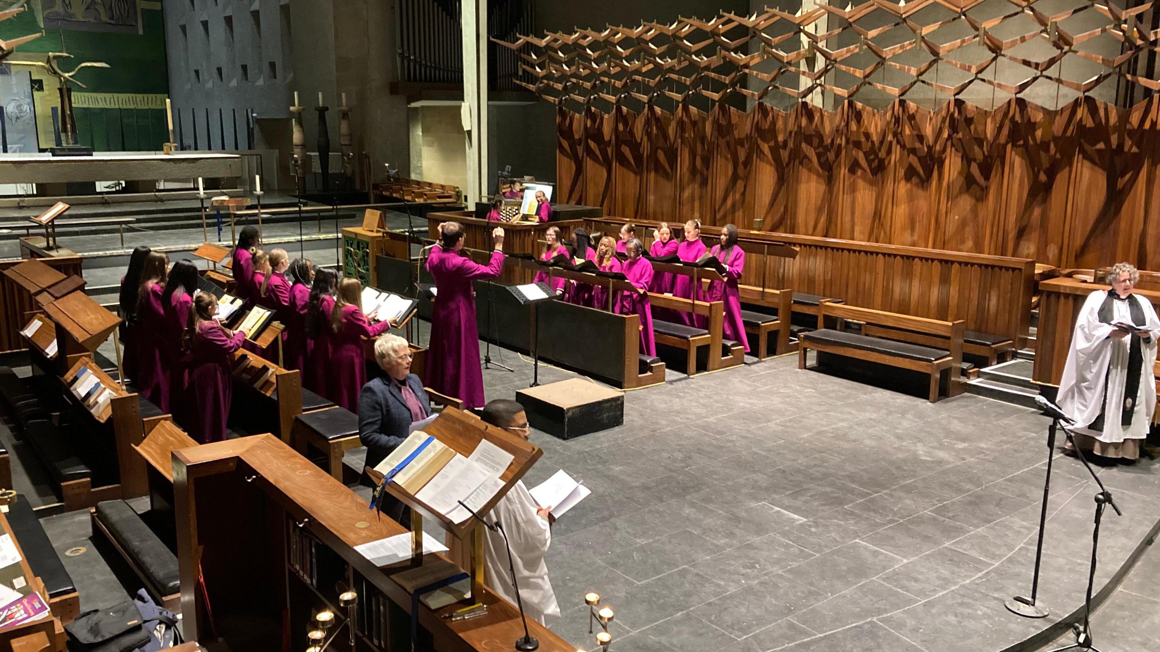 Mauve-robed choristers sing, conducted by a man in similar robes. More than a dozen are split either side of him, in a wood panelled with stone paving on the floor.