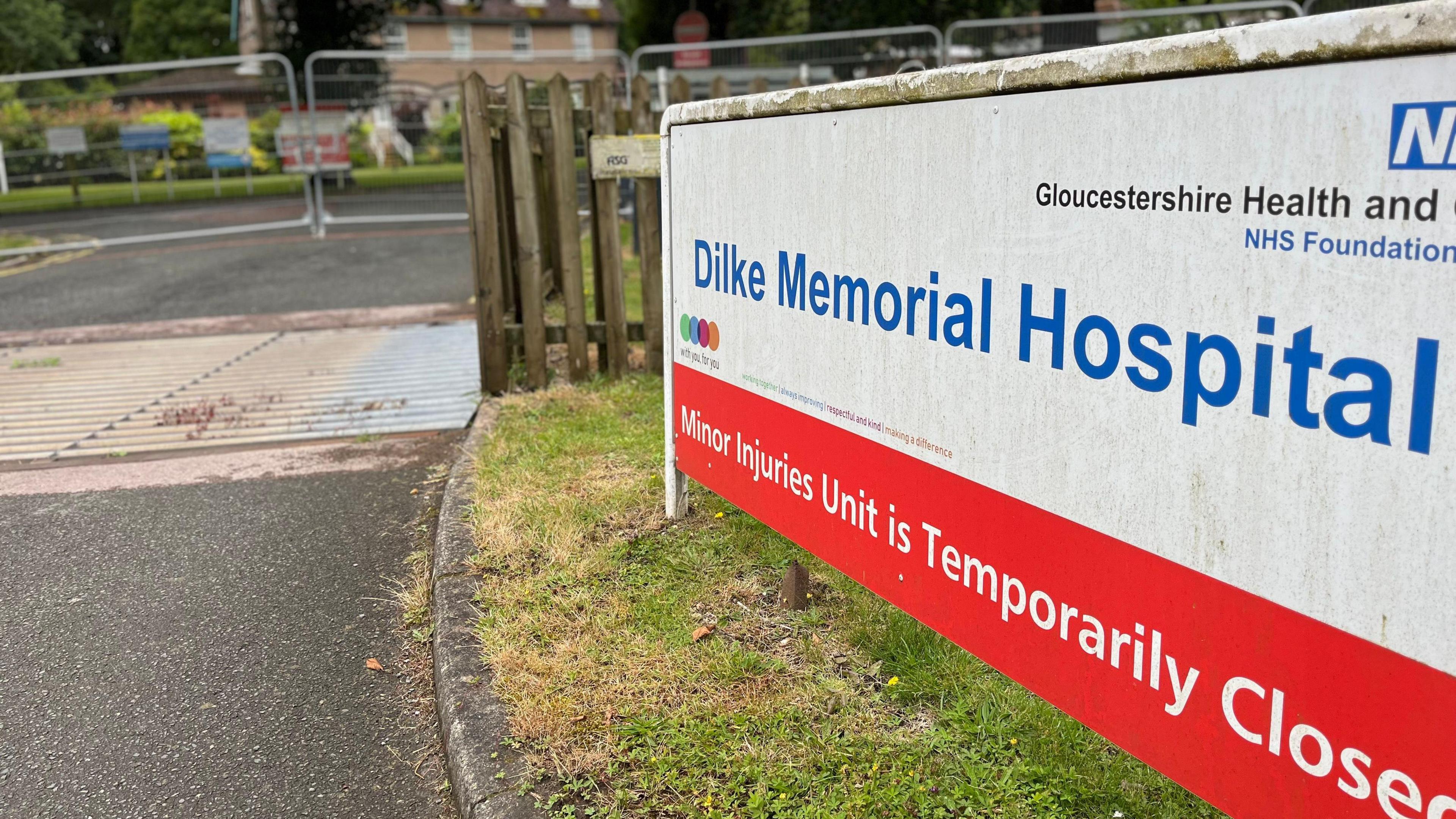 A sign bearing the name of Dilke Memorial Hospital followed by a Minor Injuries Unit is temporarily closed sign. There are barricades in the property in the background