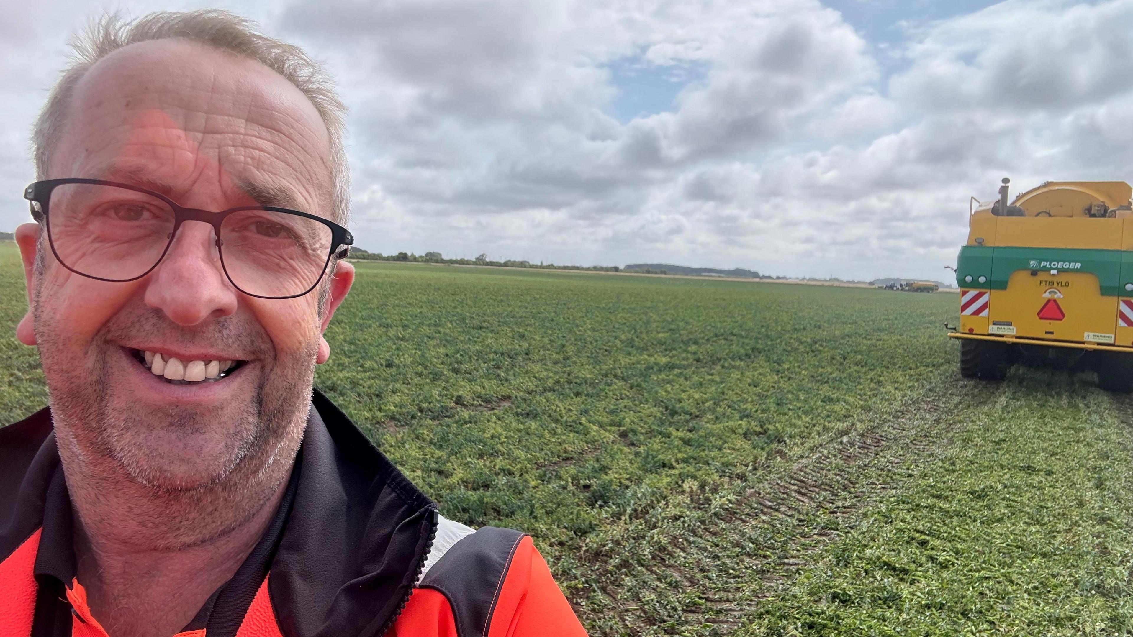 Ian has thinning grey hair and is standing in a pea field wearing an orange high-vis jacket. He is on the far left of the shot. On the far right is the rear of a yellow combine harvester