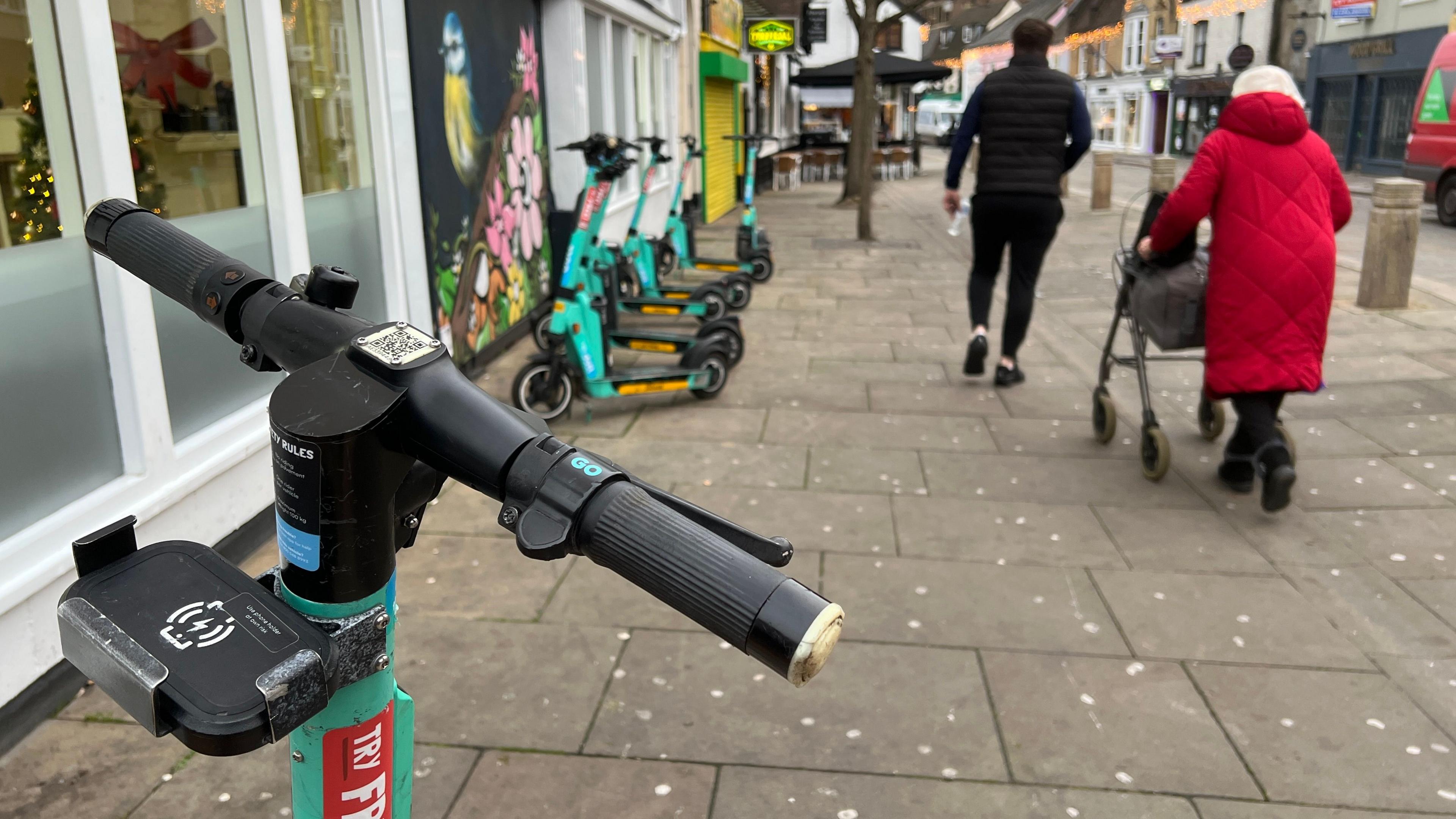 E-scooters parked on a wide pavement in a city centre with an old lady in large road coat walking past them aided with a walker.