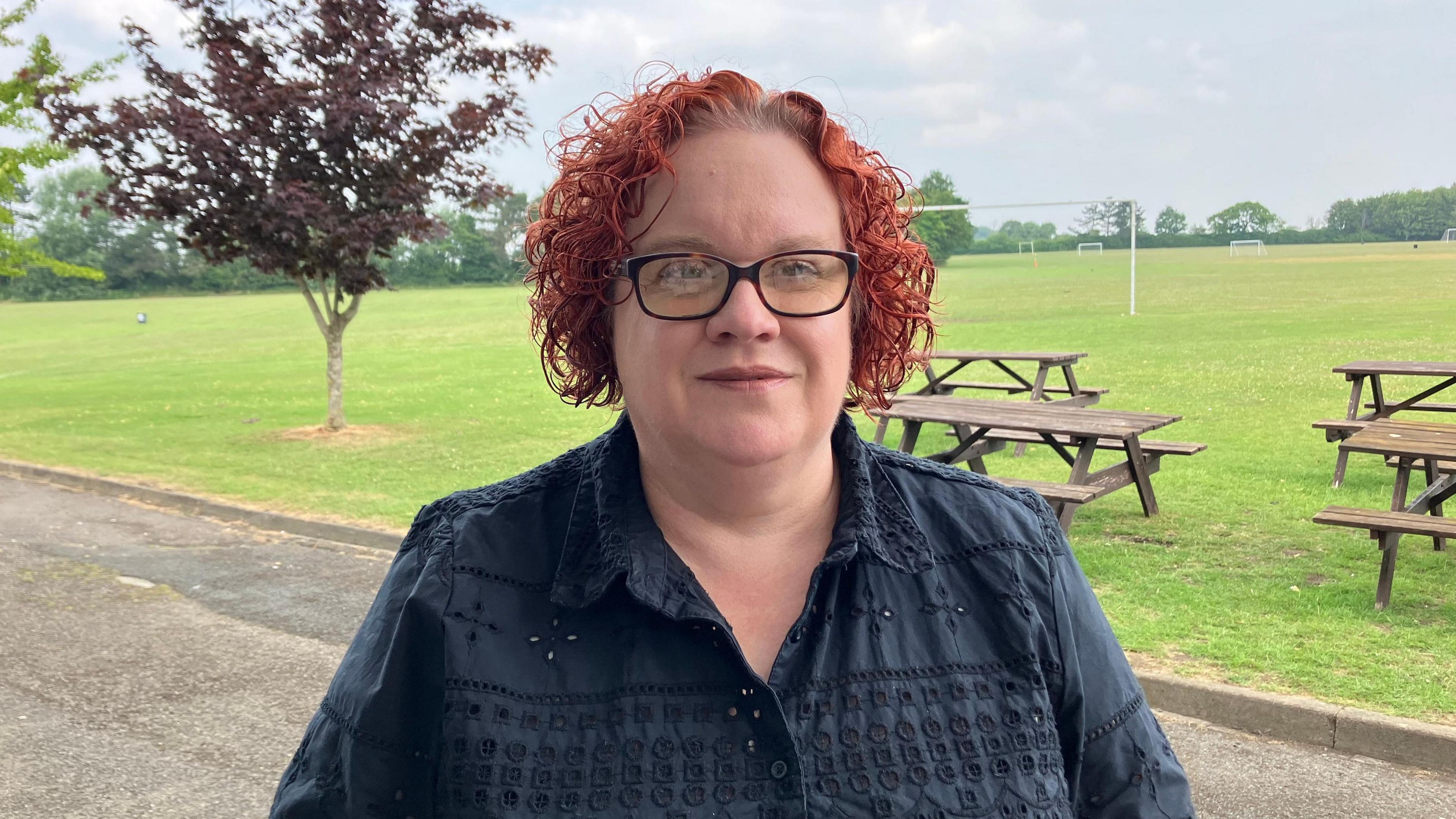 A woman standing by a playing field in a black top with red curly hair. There is a tree to the left of her head and picnic benches on the right.