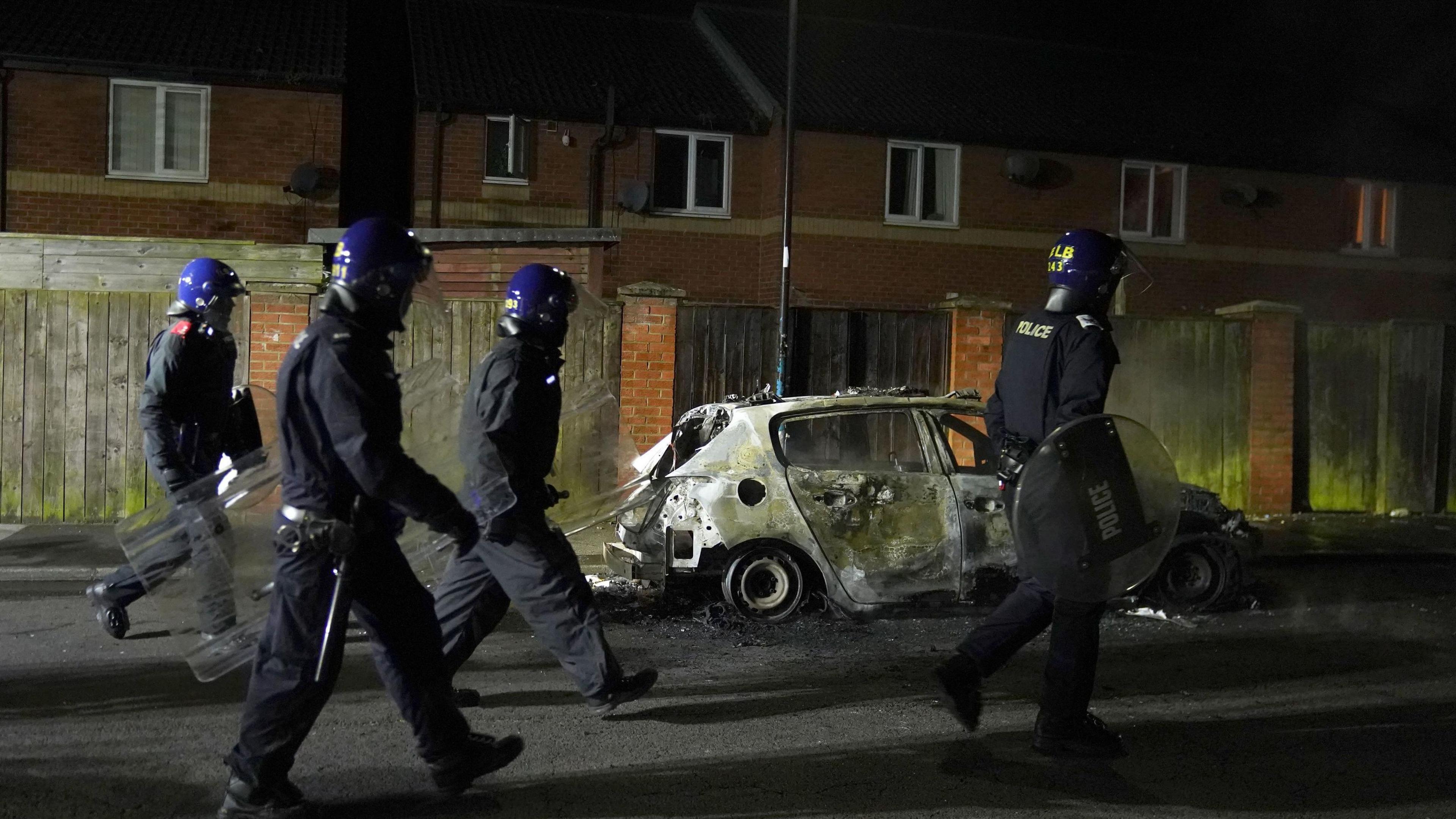 Officers walking past a burnt-out police vehicle.