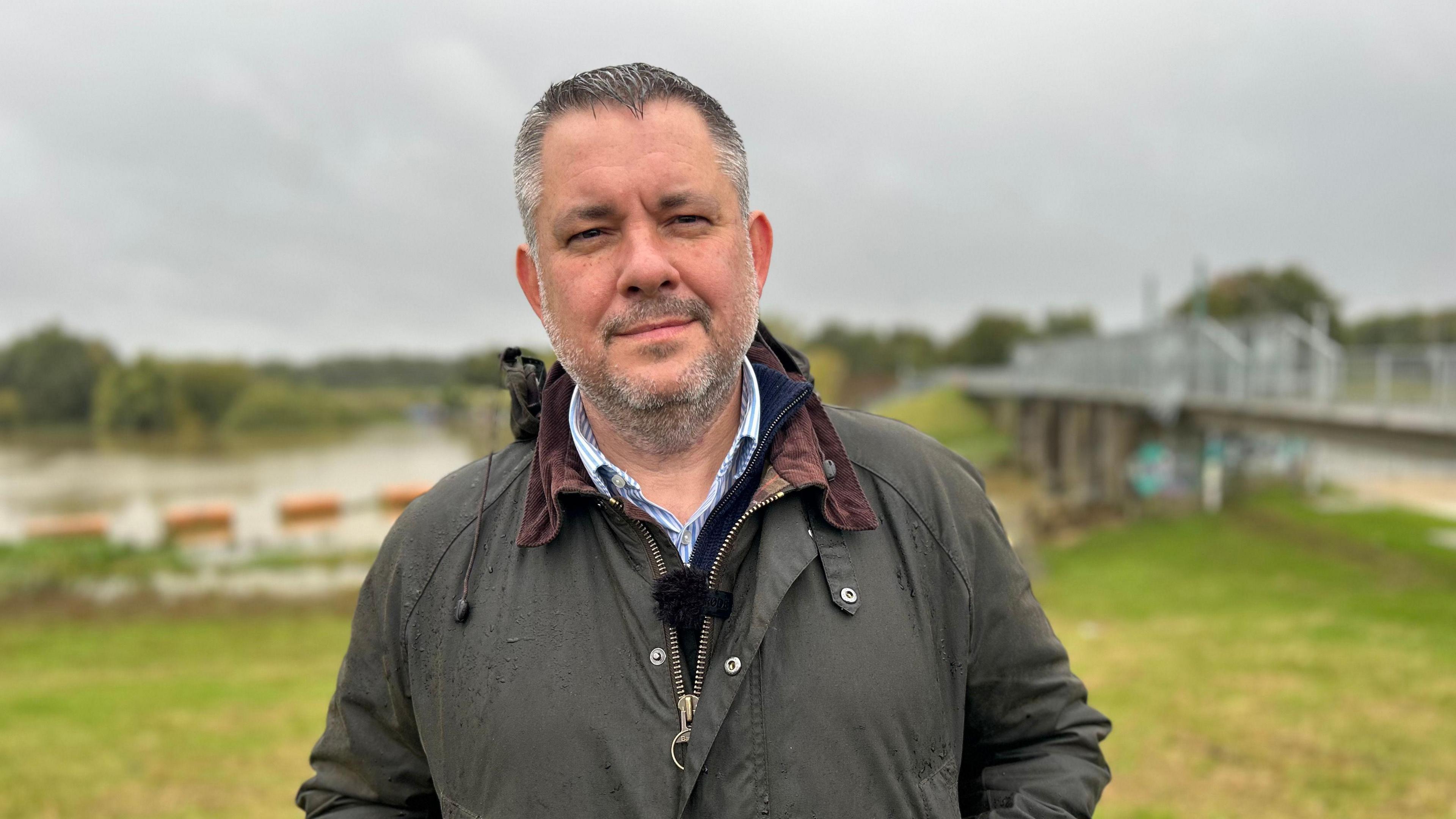 A man in an olive wax jacket stands centrally in a field smiling at the camera. There appears to be a river and a footbridge behind him.