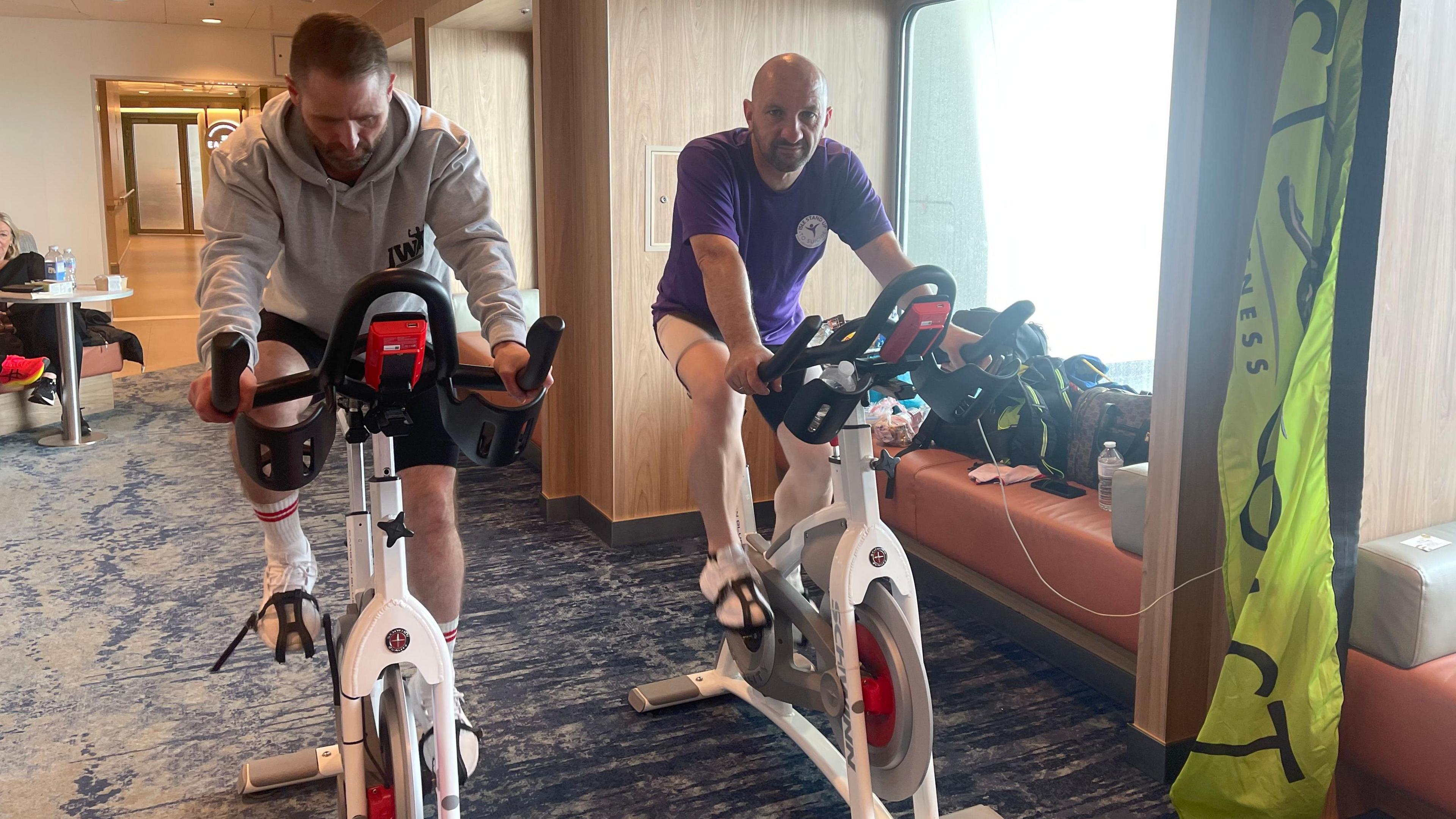 Darren, a man with short brown hair and a grey jumper and Adrian a bald man with a purple t-shirt on exercise bikes on the Manxmman.