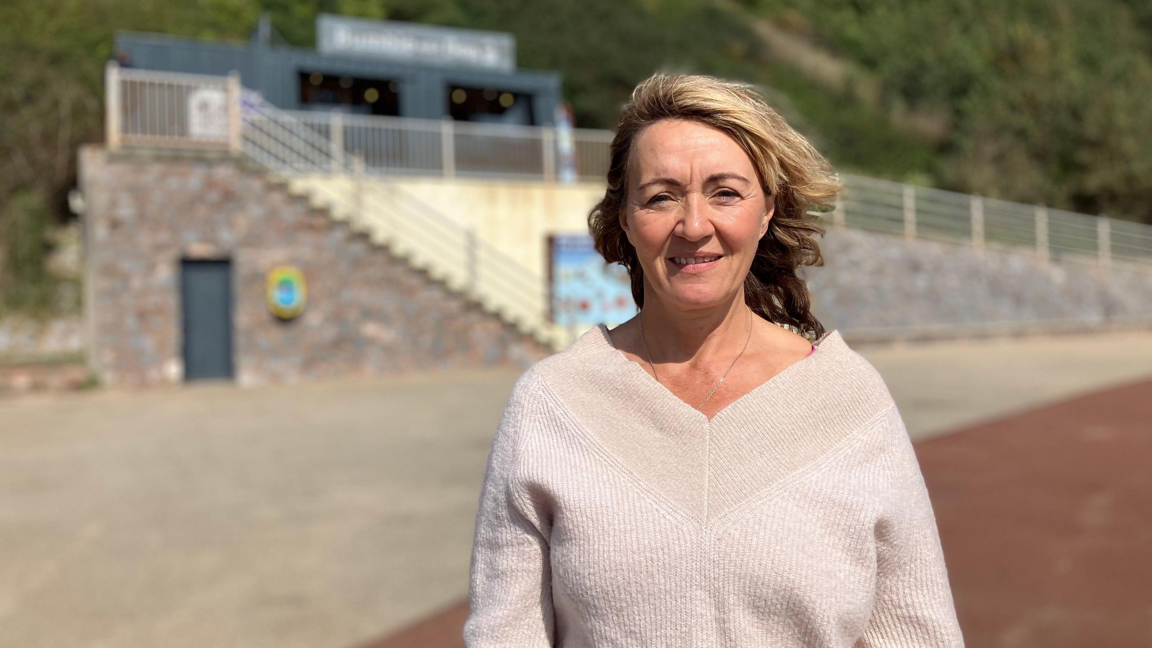 Debra Quine stood near Exmouth beach. She has blonde hair blowing in the wind and is wearing a white jumper. She is stood in front of a set of steps leading to a platform where a grey business unit is placed.