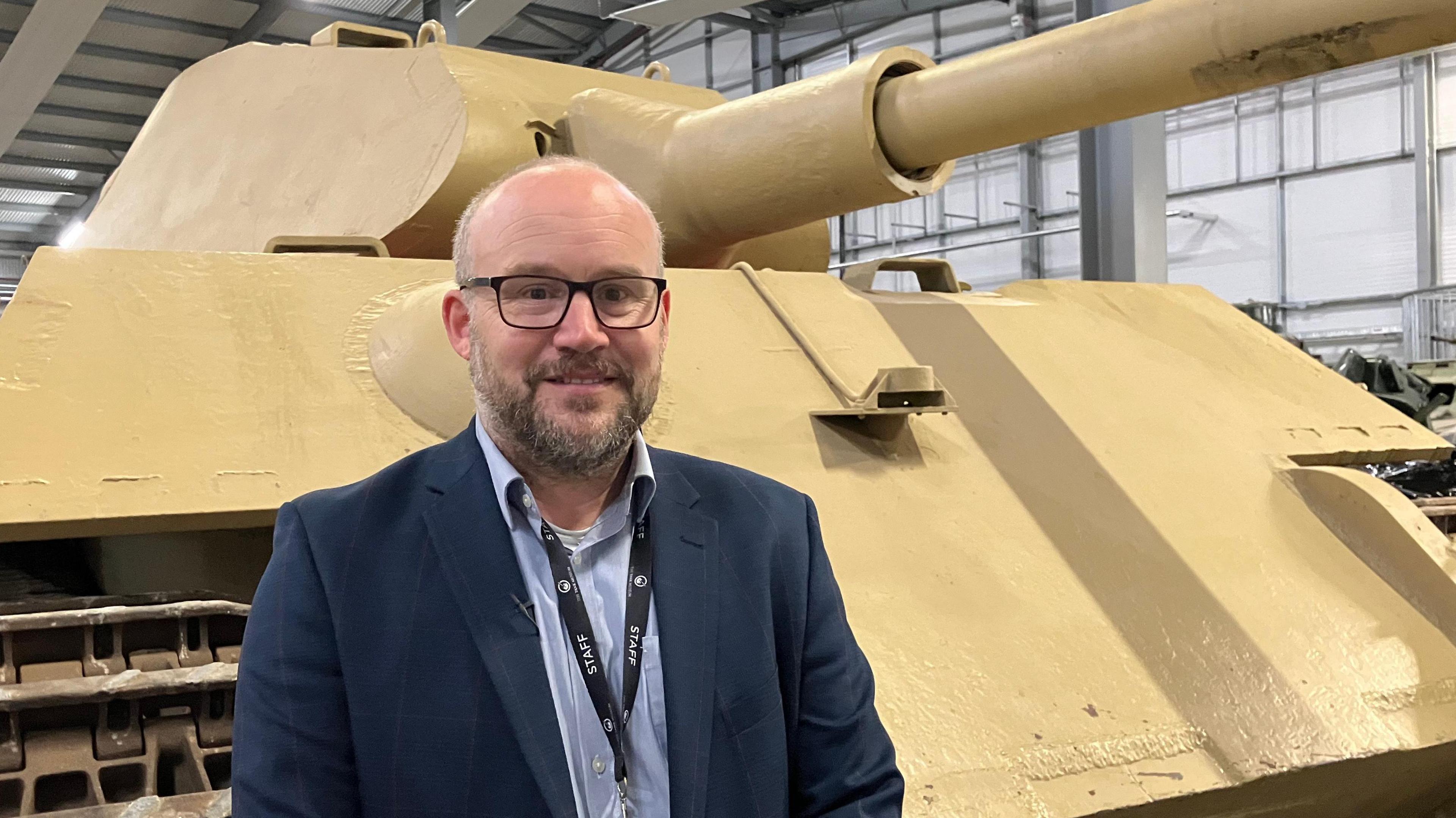 Chris van Schaardenburgh, The Tank Museum's Vehicle Collections Manager stands in front of the King Tiger tank. He is wearing a dark jacket with a light blue shirt, he has a beard and wears glasses.