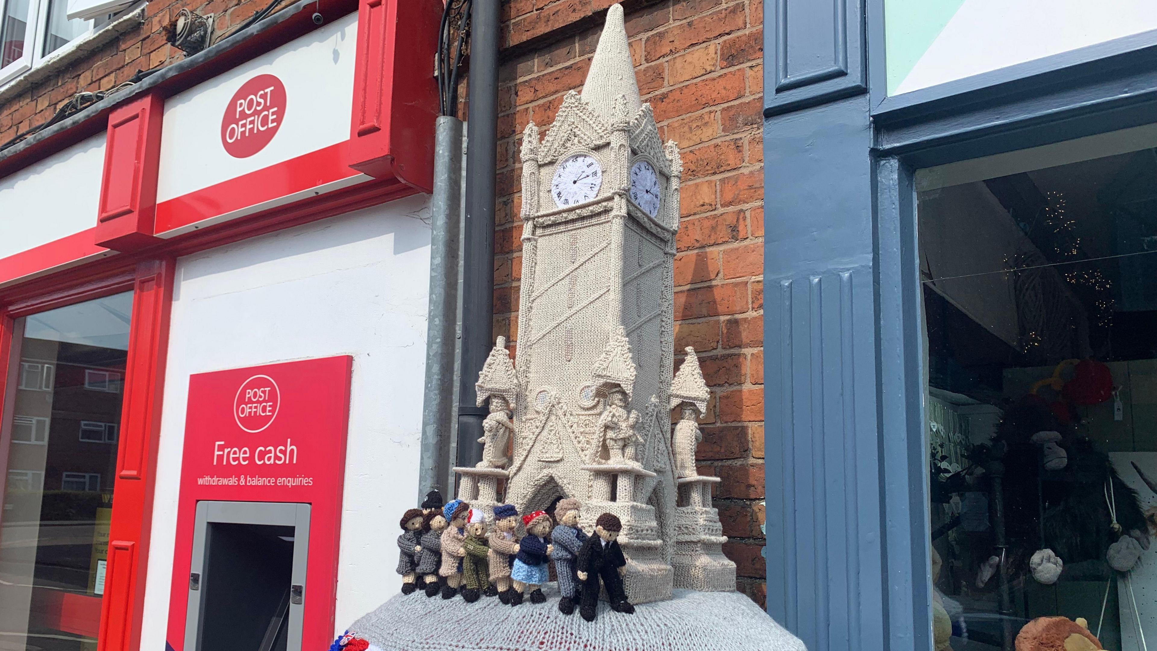 Knitted postbox topper with knitted figures doing a conga around Leicester's clock tower.