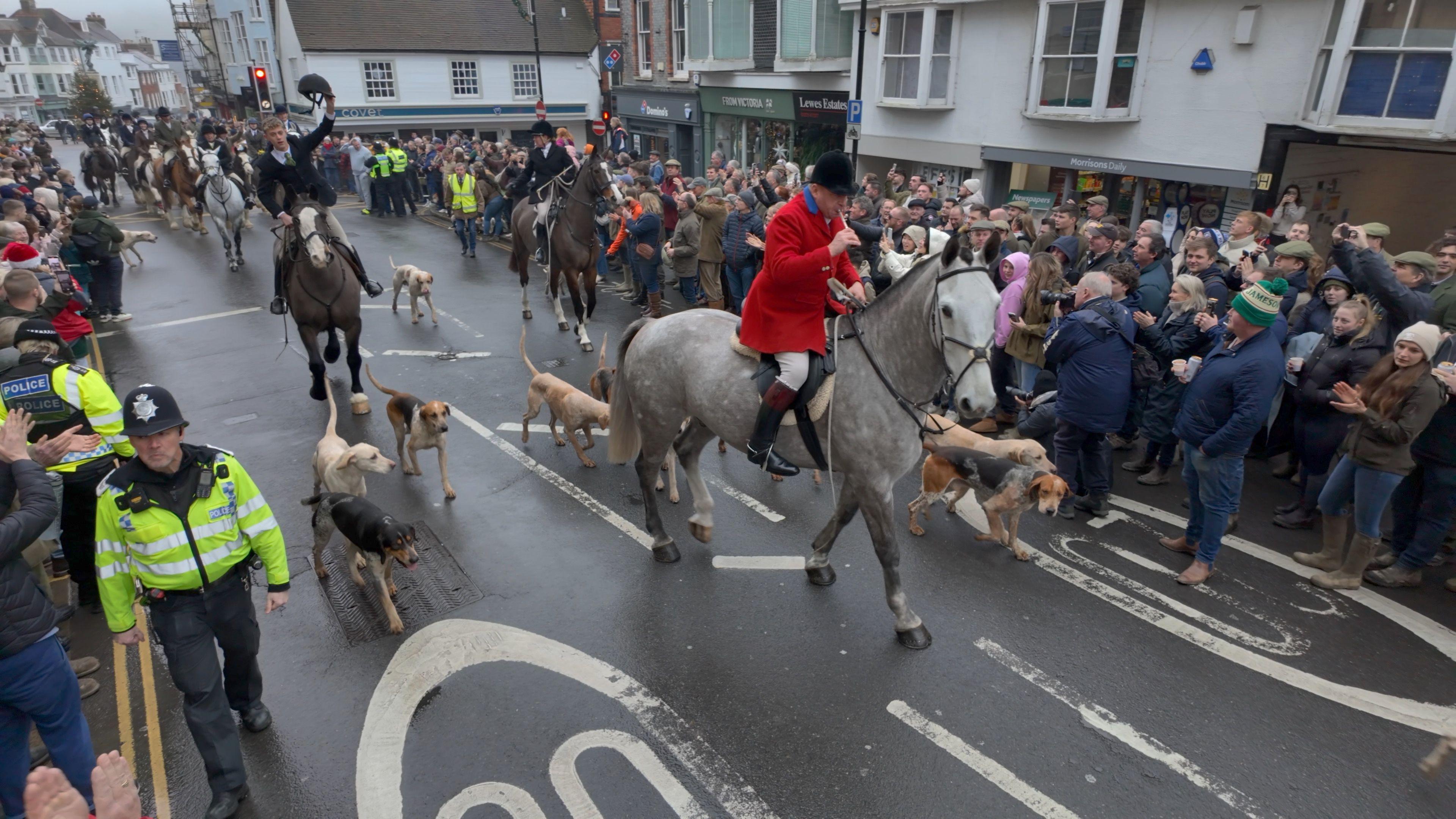 A grey horse ridden by a man in a red jacket leads other ridden horses through streets lines with police and protesters.