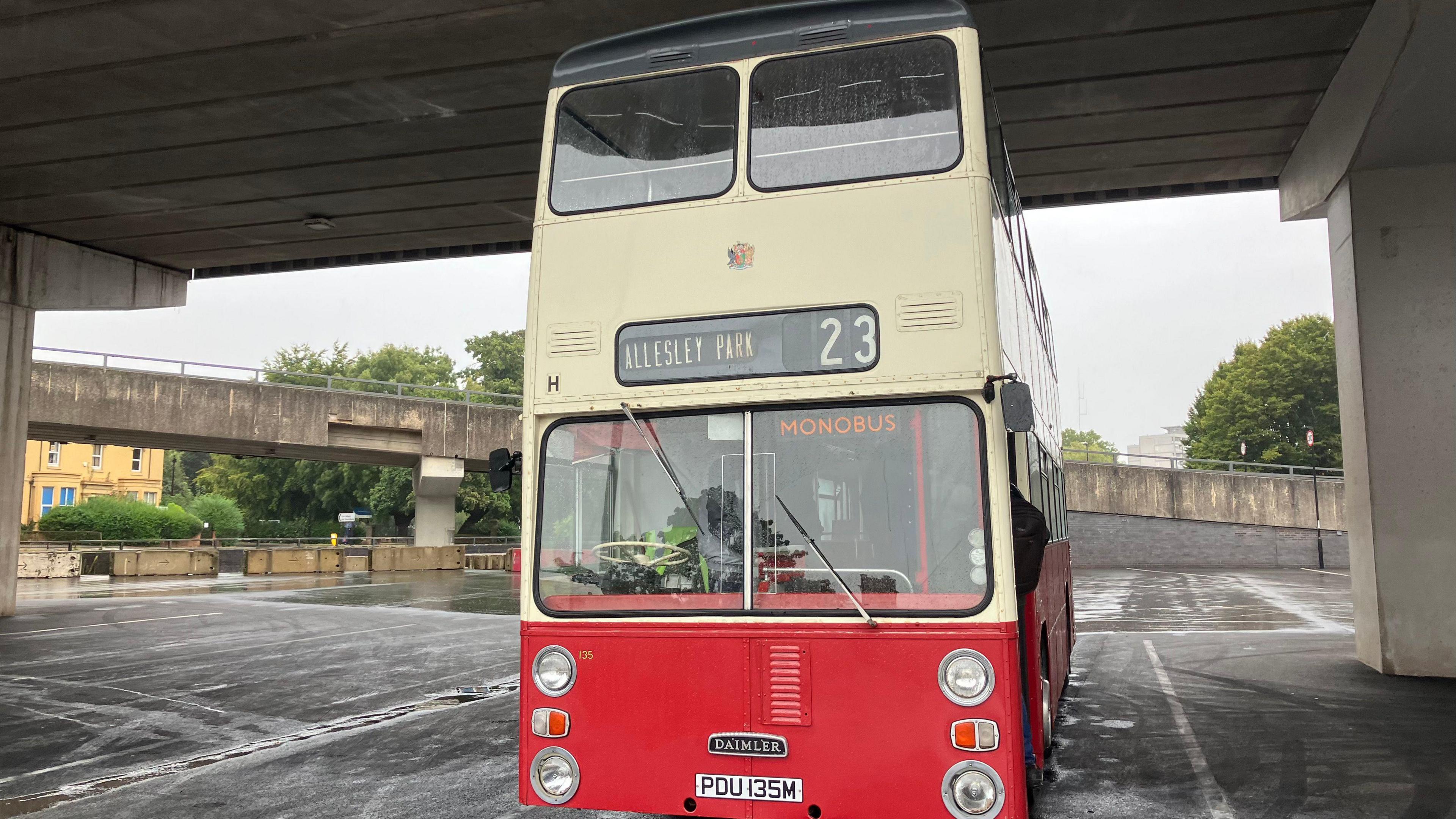 A double-decker vintage bus, with red paint and the bottom and cream at the top, in a car park under a bridge. The bus sign says 'Allesley Park 23'.