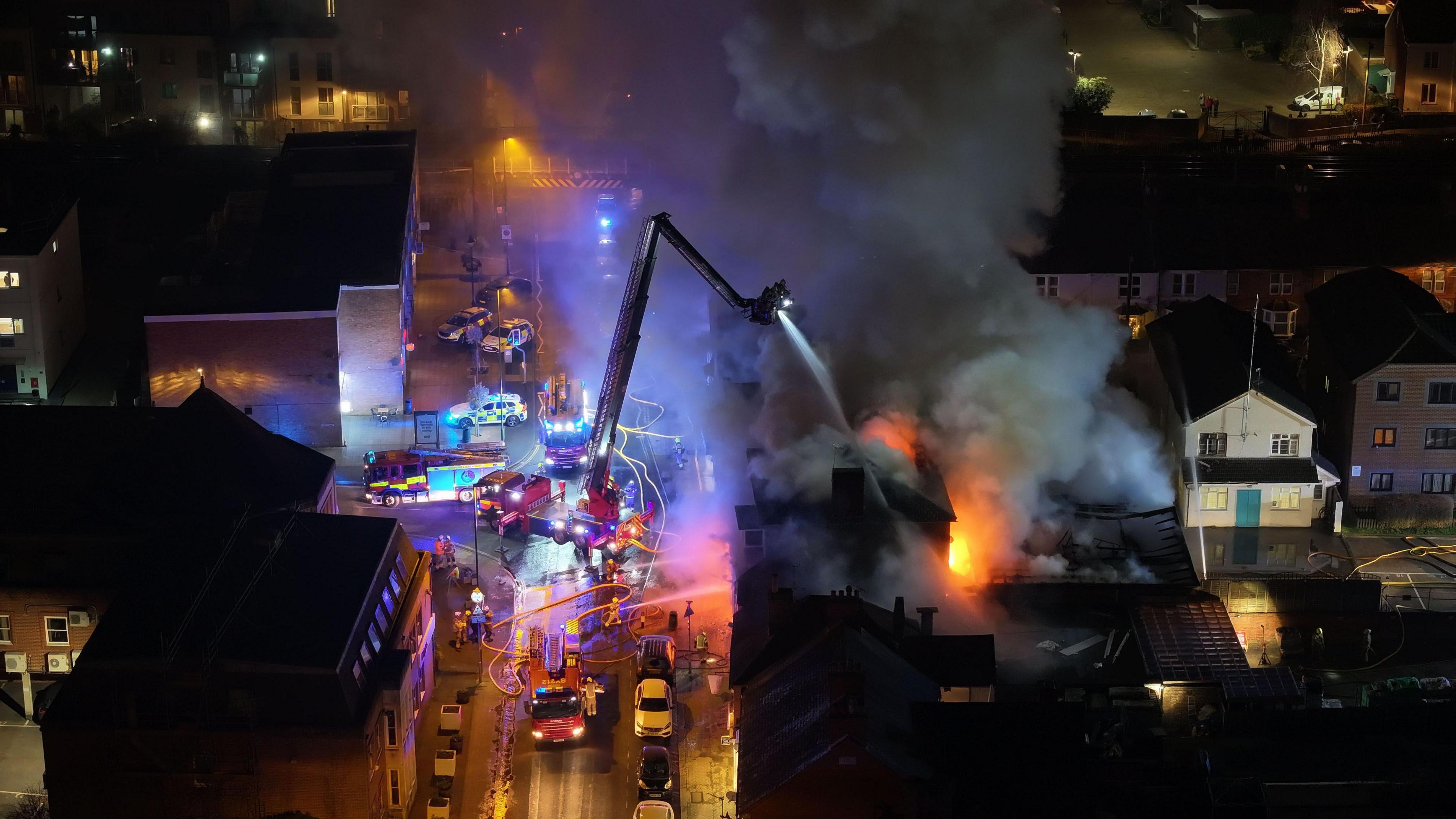 Huge plumes of black smoke billow up from the rooftop of the building and orange flames are glowing out of the front and top of the building. Firefighters and police are at the scene. Firefighters are on the road spraying the fire and a large hydraulic platform is spraying the fire from above