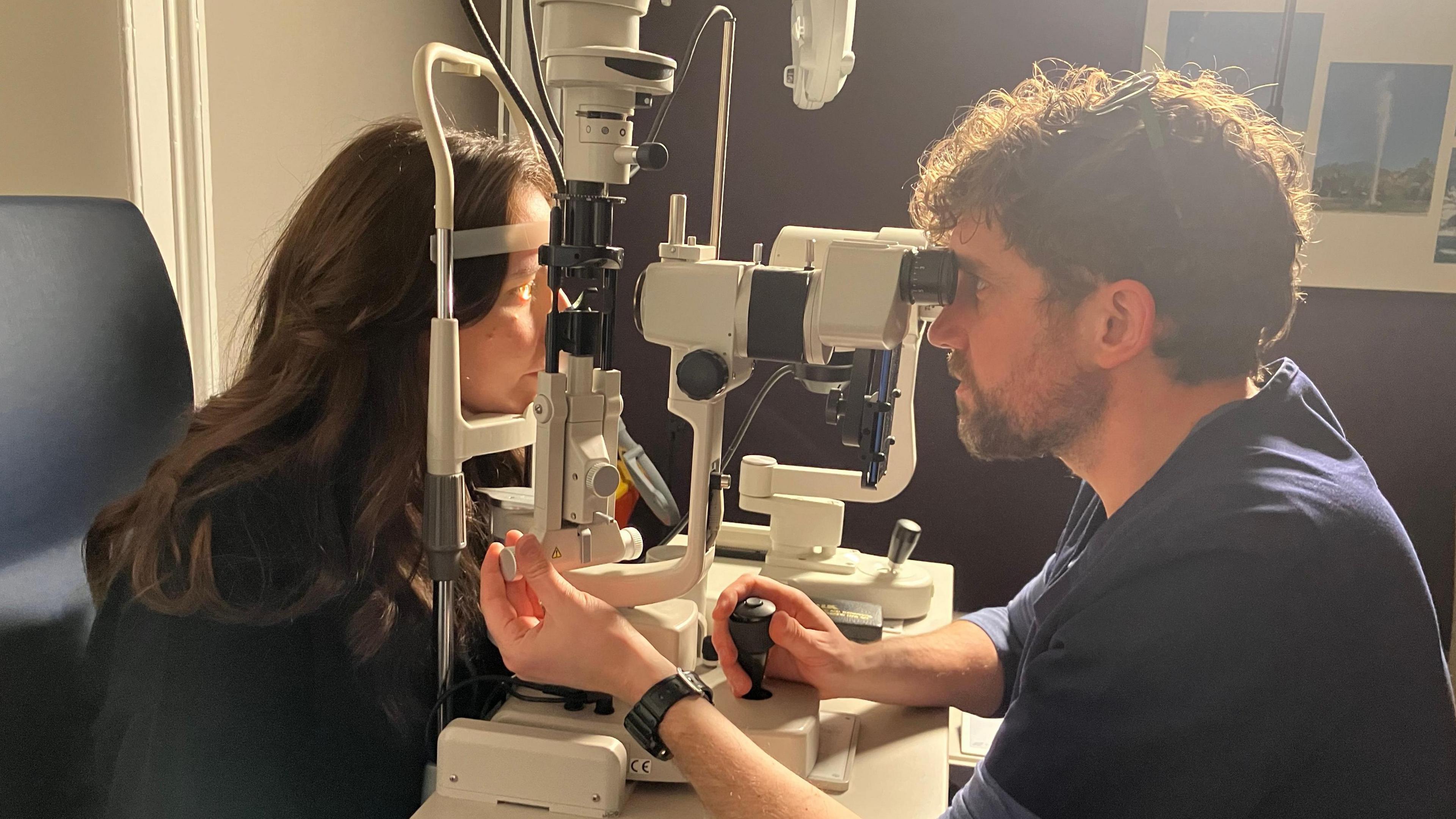 Optometrist Ian Cameron giving a woman an eye examination through a piece of specialised equipment. He is looking into the machinery on one side as she rests her chin on the either side as he looks into her eye. One of her eyes is lit up.