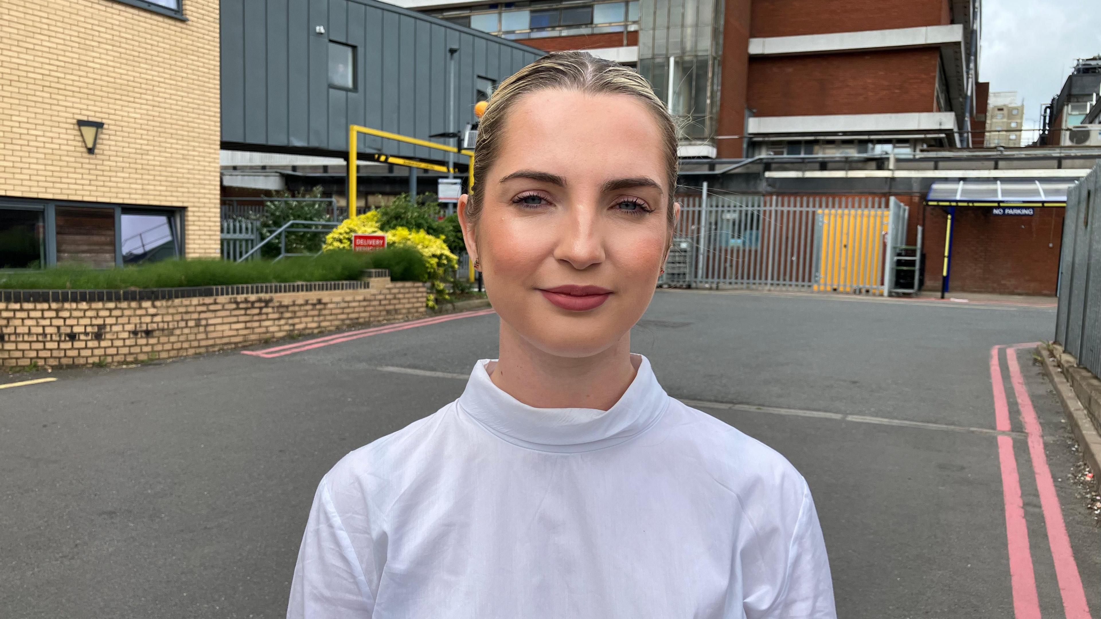 Rosie Wrighting stands outside the hospital in a car park. She has blonde,  hair which is tied back and is wearing a white roll-neck top. She has a serious facial expression. 