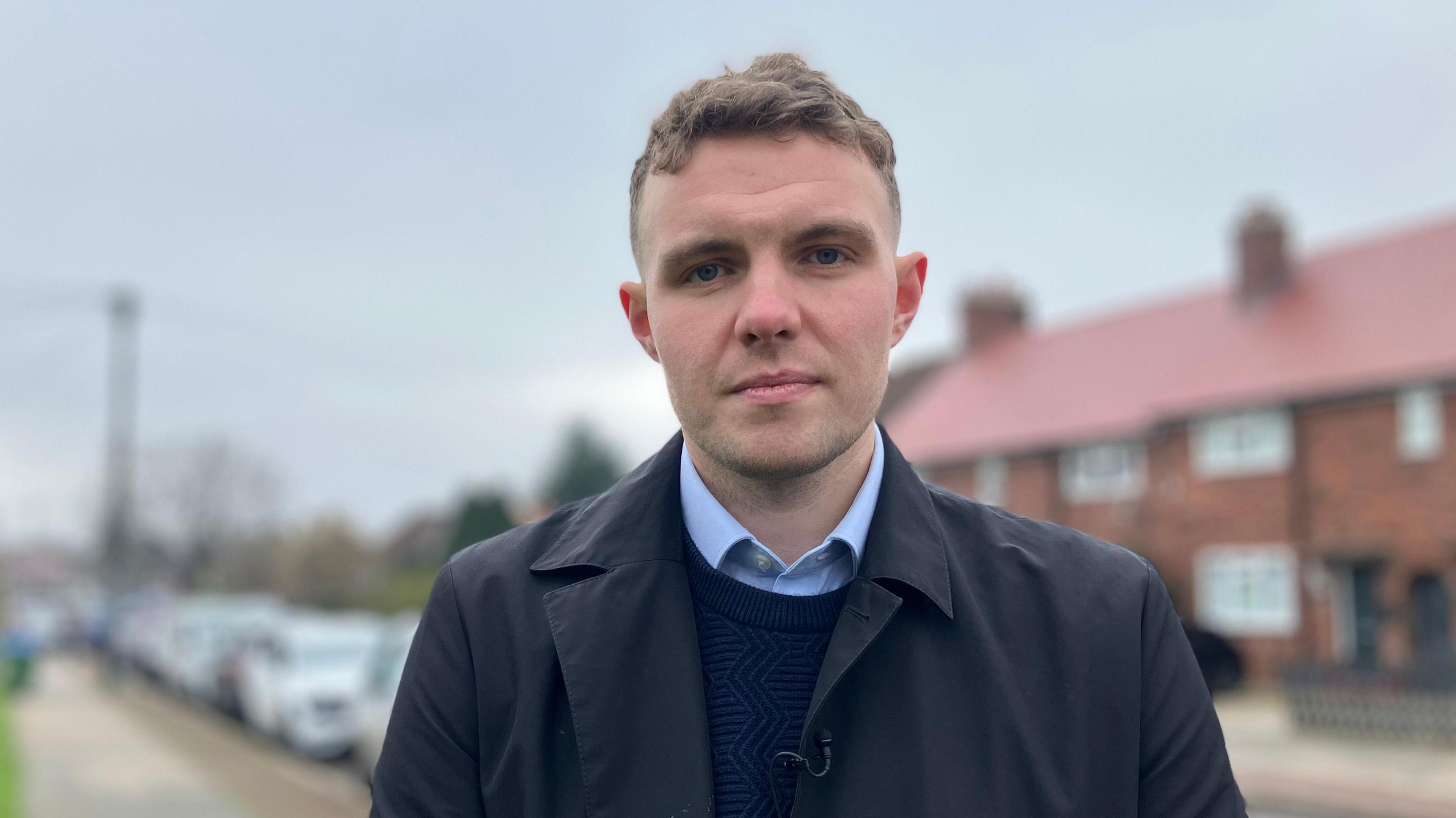 Charlie Davis is seen facing forward. He is standing outside on a residential street wearing a blue jumper and shit with a navy coat. Red-bricked houses can be seen in the background and a row of parked cars.