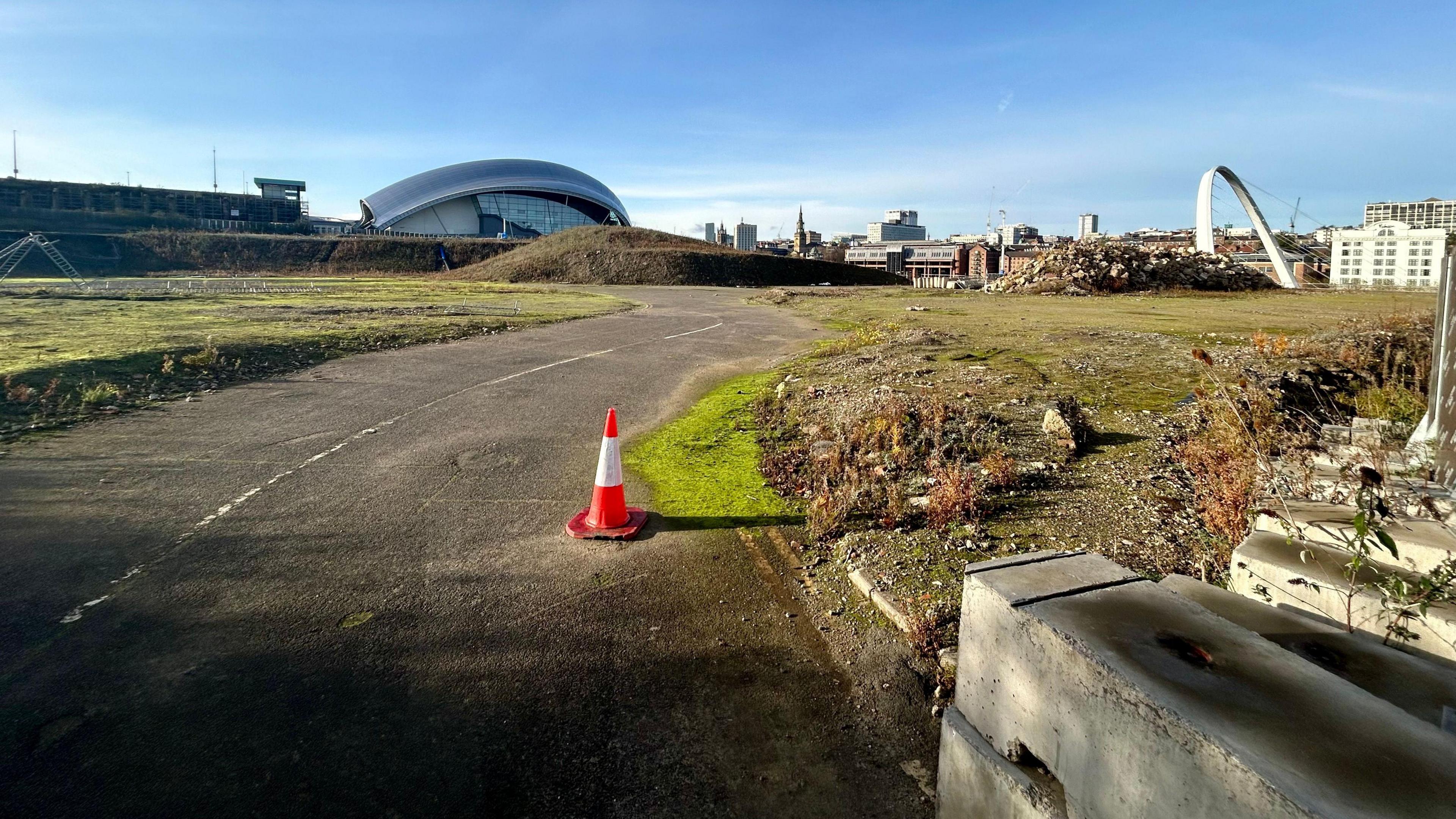 A single traffic cone sits on a road through empty scrubland where the proposed Sage arena and conference centre on Gateshead Quayside is due to be built. The Newcastle skyline can be seen in the distance.