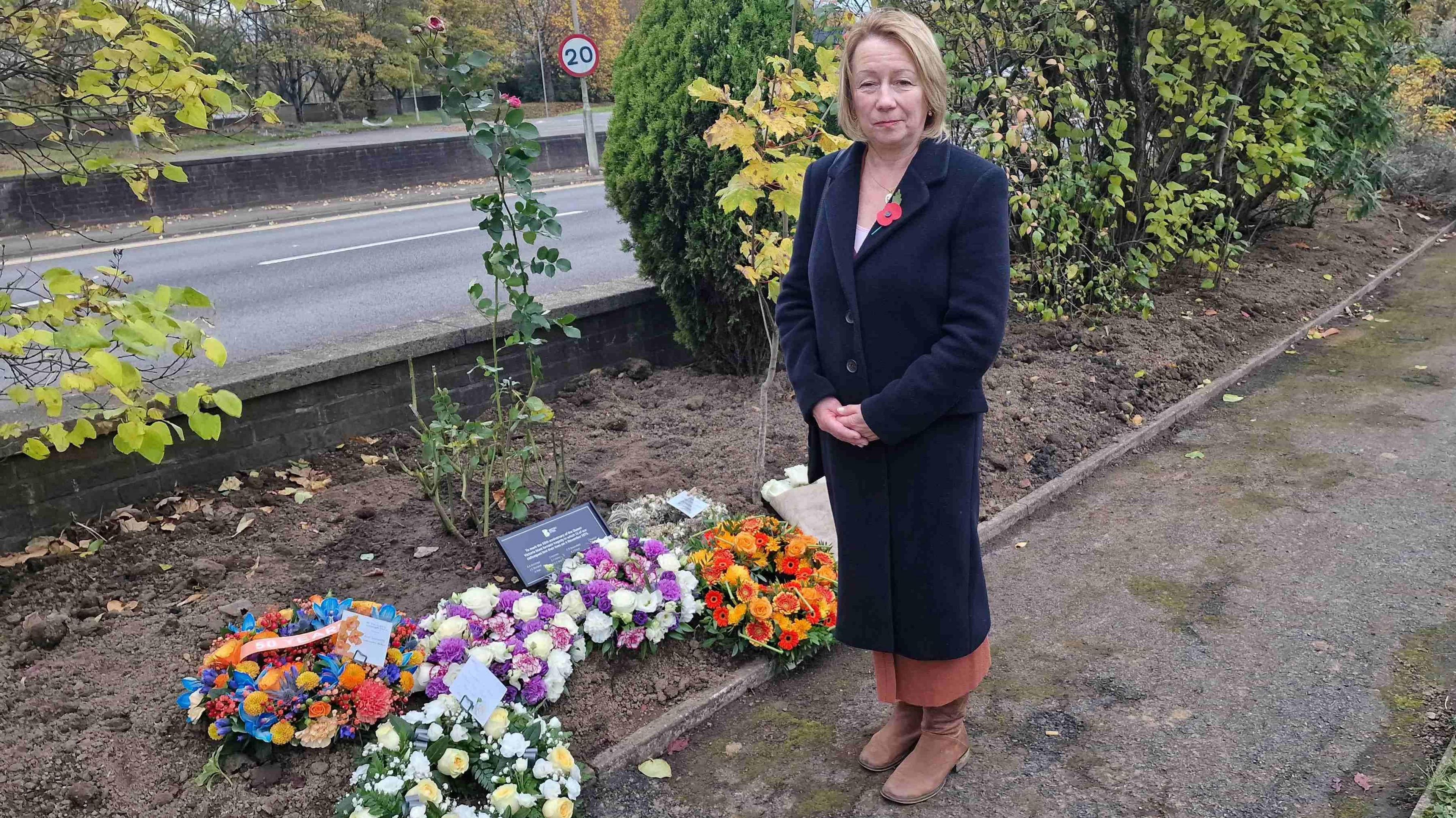 Woman with shoulder length blonde hair wearing a dark coat, white top and peach skirt with brown shoes. She is wearing a poppy. She is stood next to wreaths laid next to a plaque. There are bushes and a road.