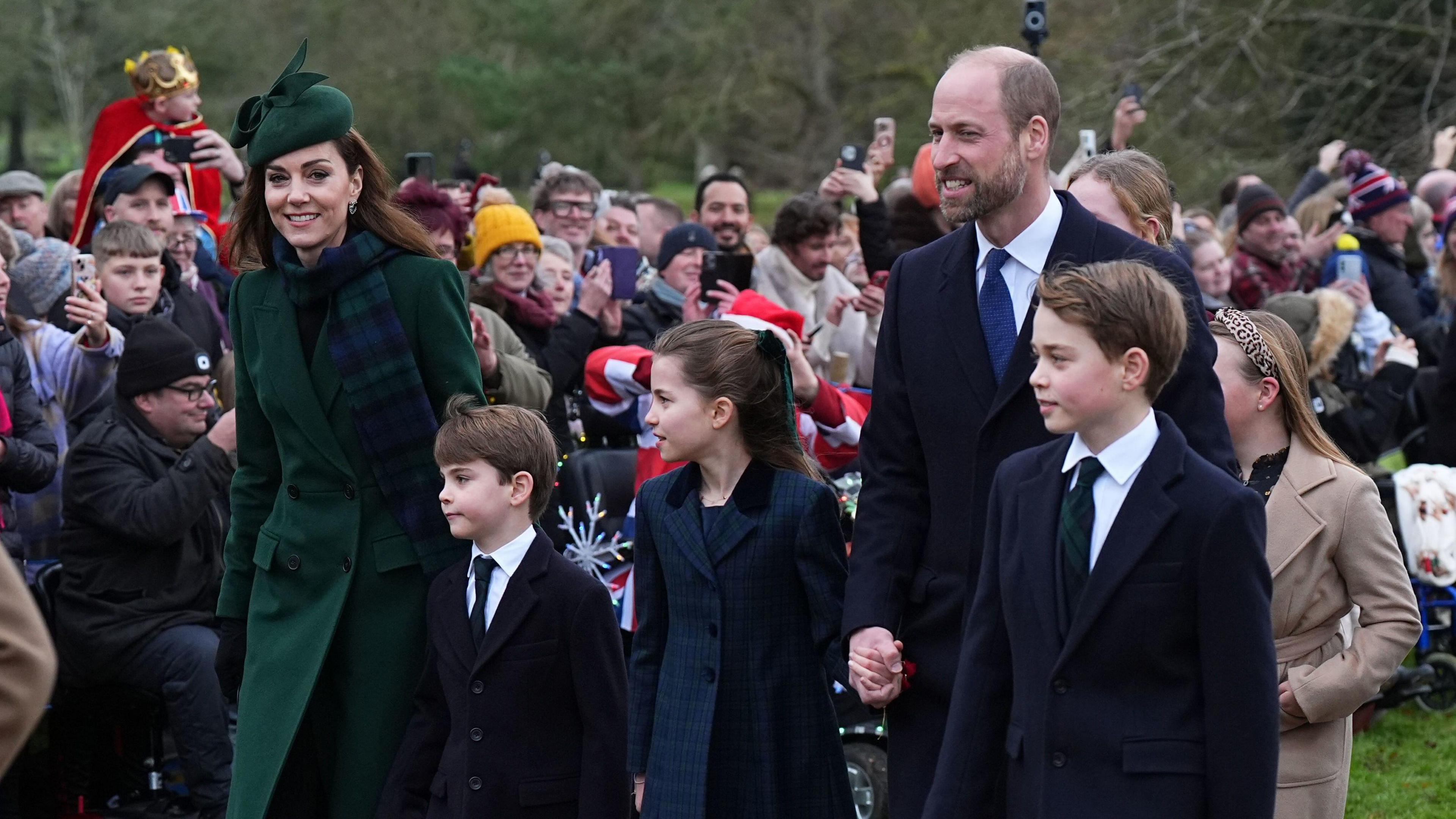 The Princess of Wales wearing a matching green hat and coat with tartan scarf accompanied by Prince William their sons George, and are wearing dark jackets with shirts and ties and her daughter Charlotte who is wearing a blue coat. 