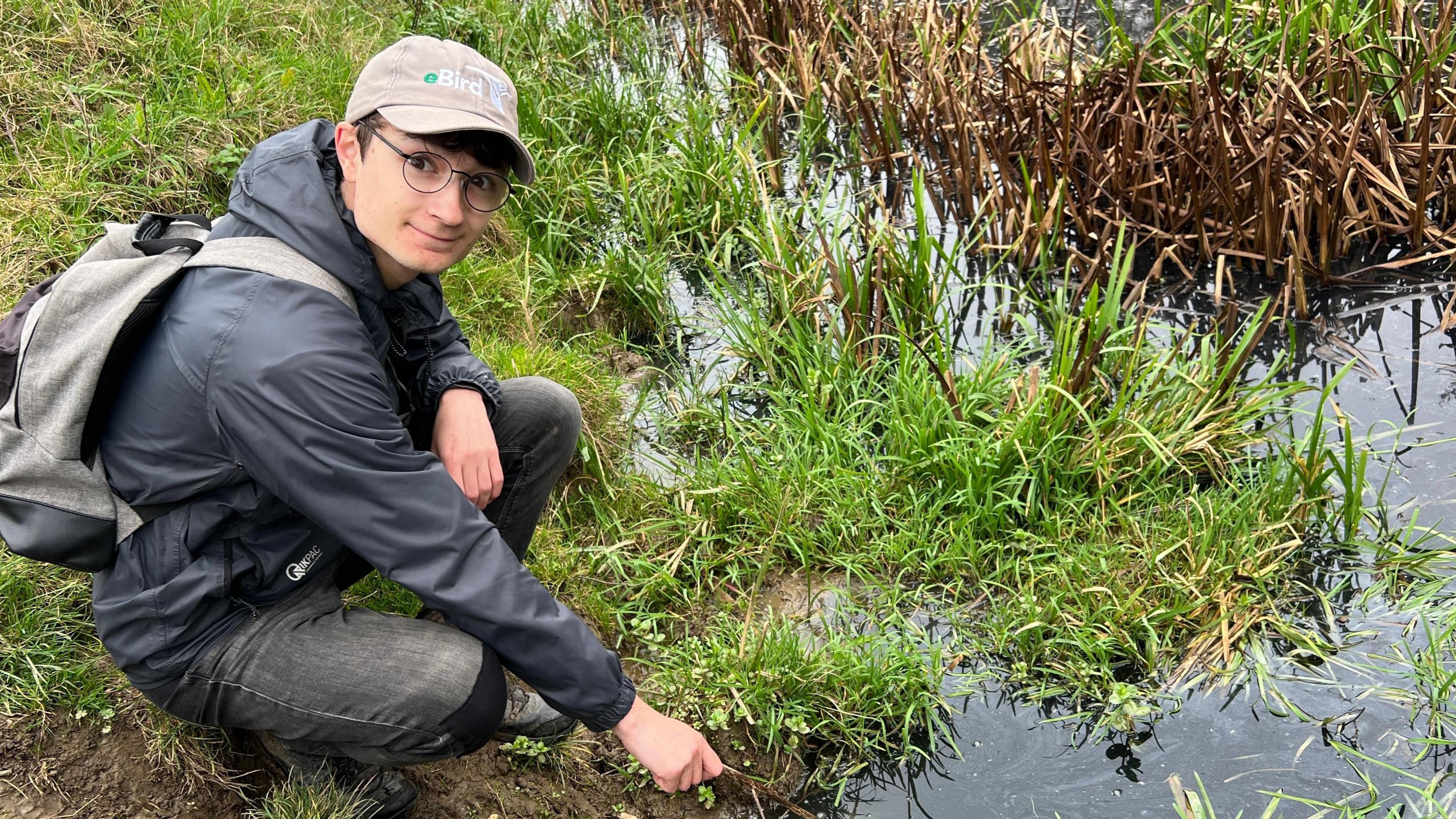 Owen Tattersall crouches down on a grassy bank and points to black water in Barmston Drain. He wears a dark-grey jacket, grey jeans, a backpack, a light brown cap and glasses. 