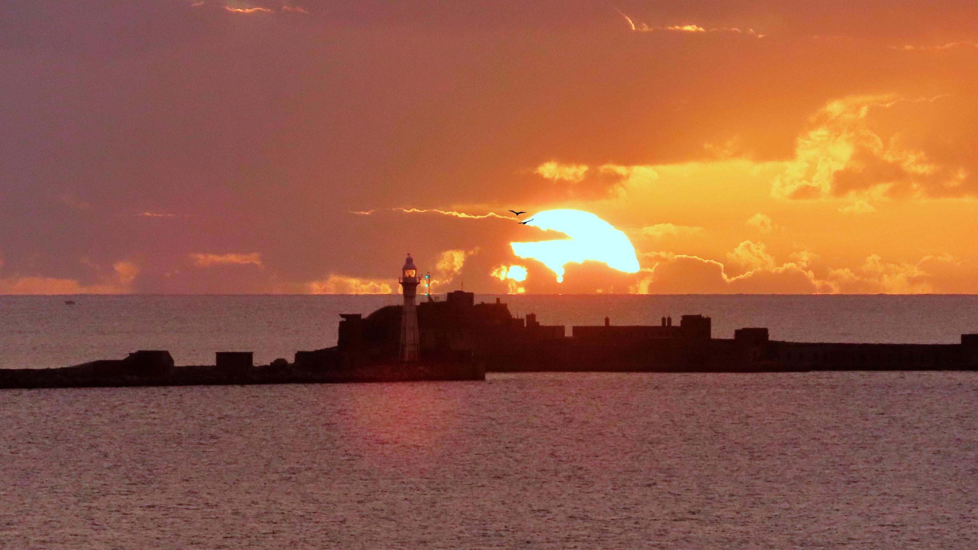 The sun rises above the sea horizon with clouds dispersing enough to clearly see  it. In front is the entrance to Weymouth Harbour and the flat calm see surrounding it.