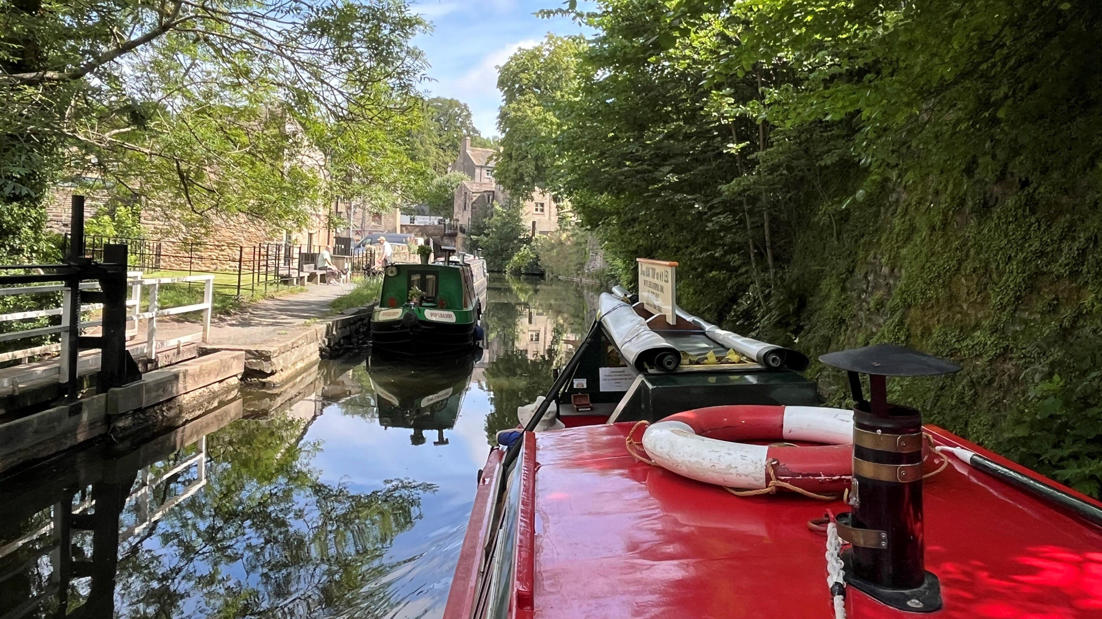 The picture has been taken onboard a red narrow boat cruising along the canal. You can see a red roof with a chimney and red and white life ring. A green narrow boat is in the distance.
