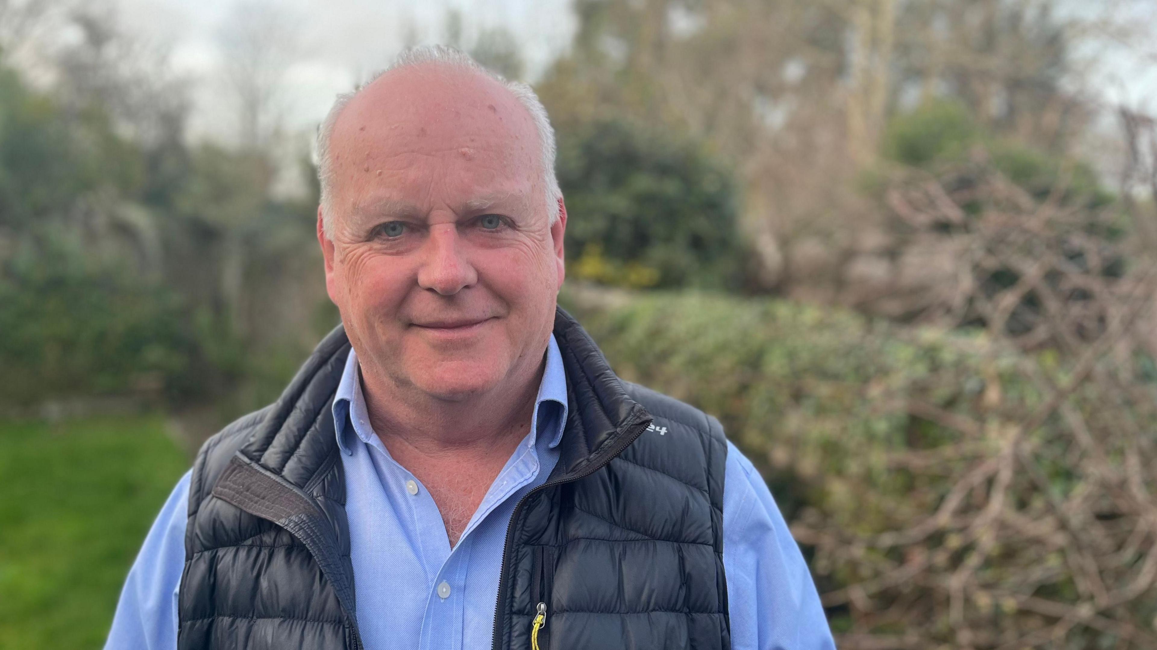 Paul Arnott, deputy leader of Devon County Council, wearing a light blue shirt and a blue gilet, standing in front of a hedgerow.