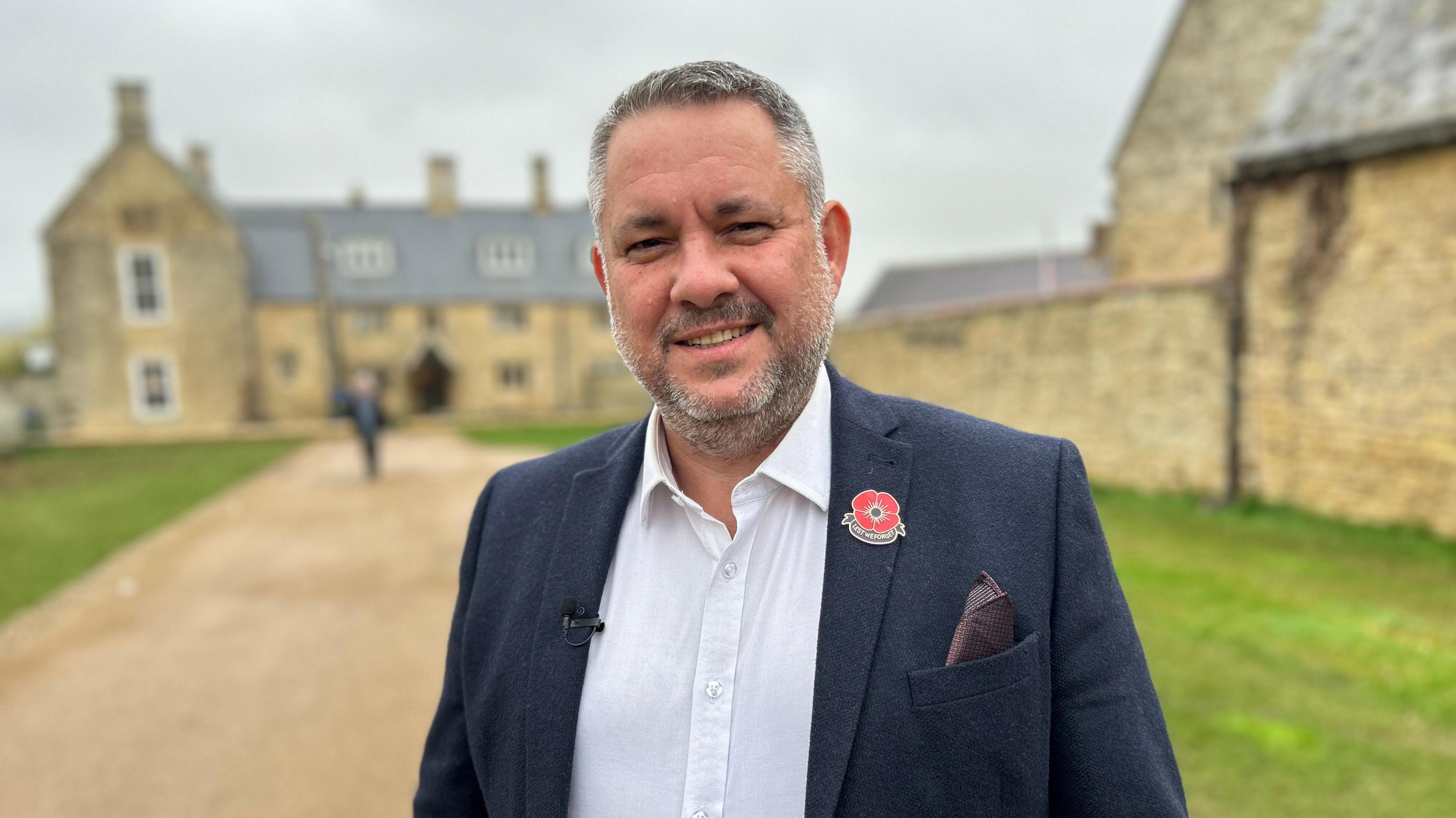 Jason Smithers wearing a navy blazer and a white shirt, standing in front of an abbey building that is blurred out. 