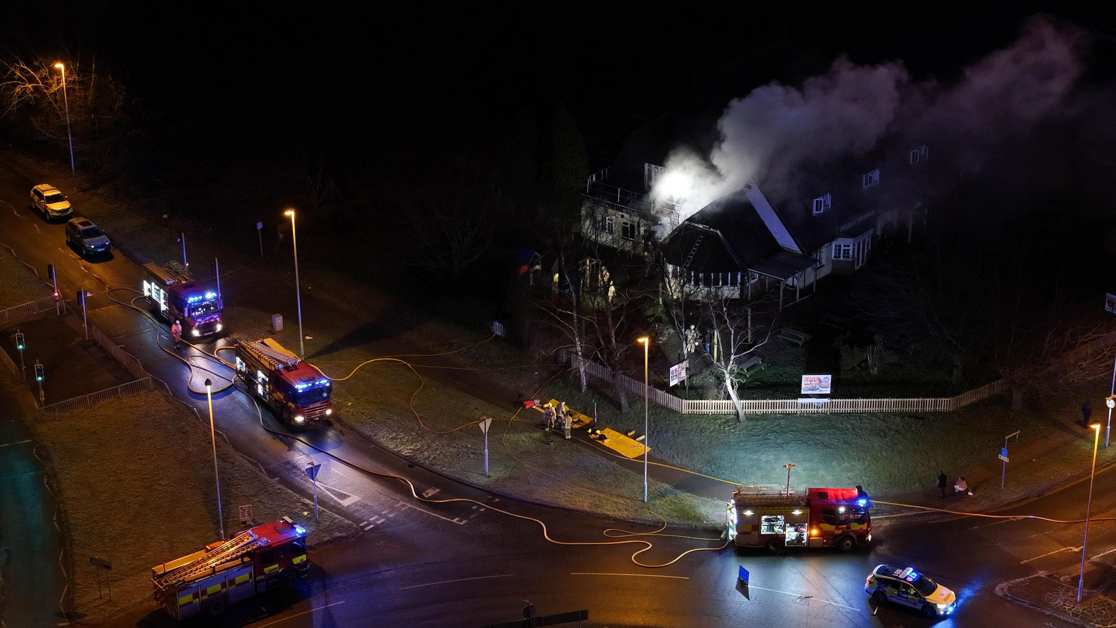 An aerial shot shows four fire engines parked at the side of the road and a large pub building is seen with smoke coming out its roof. It is dark and the vehicles have their lights on and there are fire hoses across the road and pavement