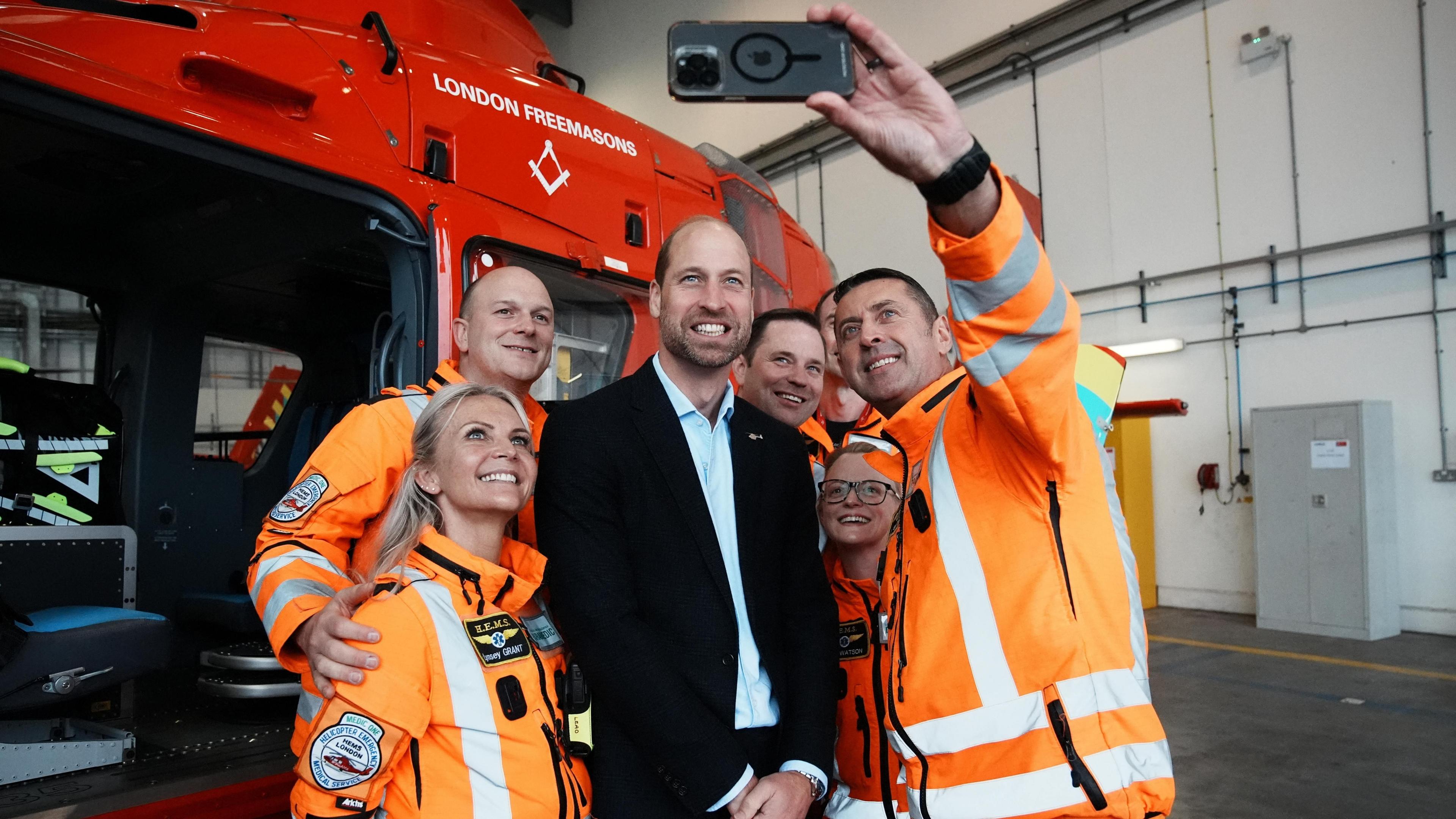 Six members of the London Air Ambulance helicopter crew, dressed in high-visibility orange and white coats pose for a selfie photograph with Prince William who is smiling and dressed in a black jacket with white shirt. In the background is a red helicopter. 