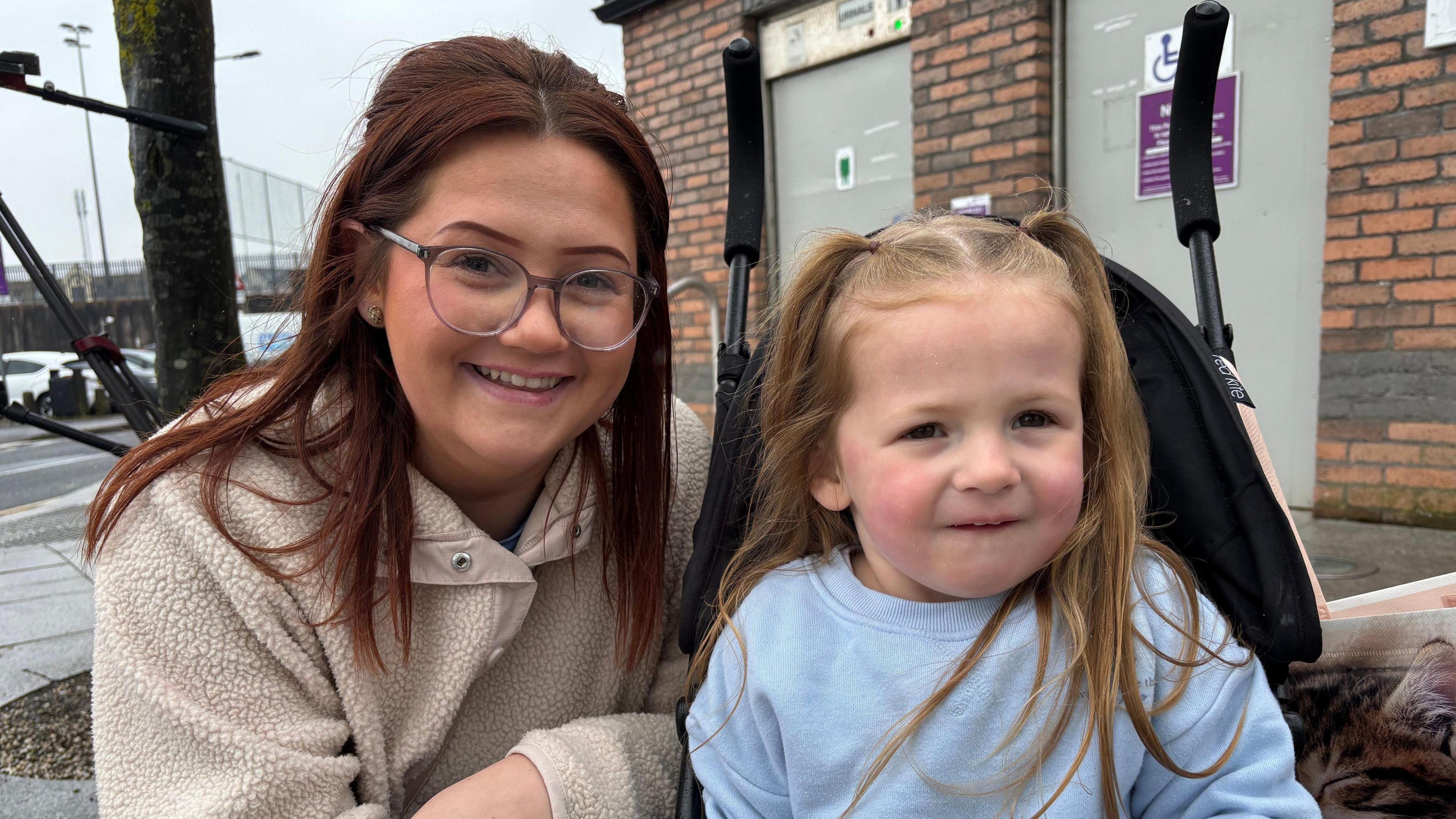 Demi Whelan and her daughter Tilly. Demi has long, brown hair and wears glasses and a cream fleece. Tilly has long, blonde hair in pigtails and wears a light blue jumper as she sits in a pram. Demi is crouched down next to her.