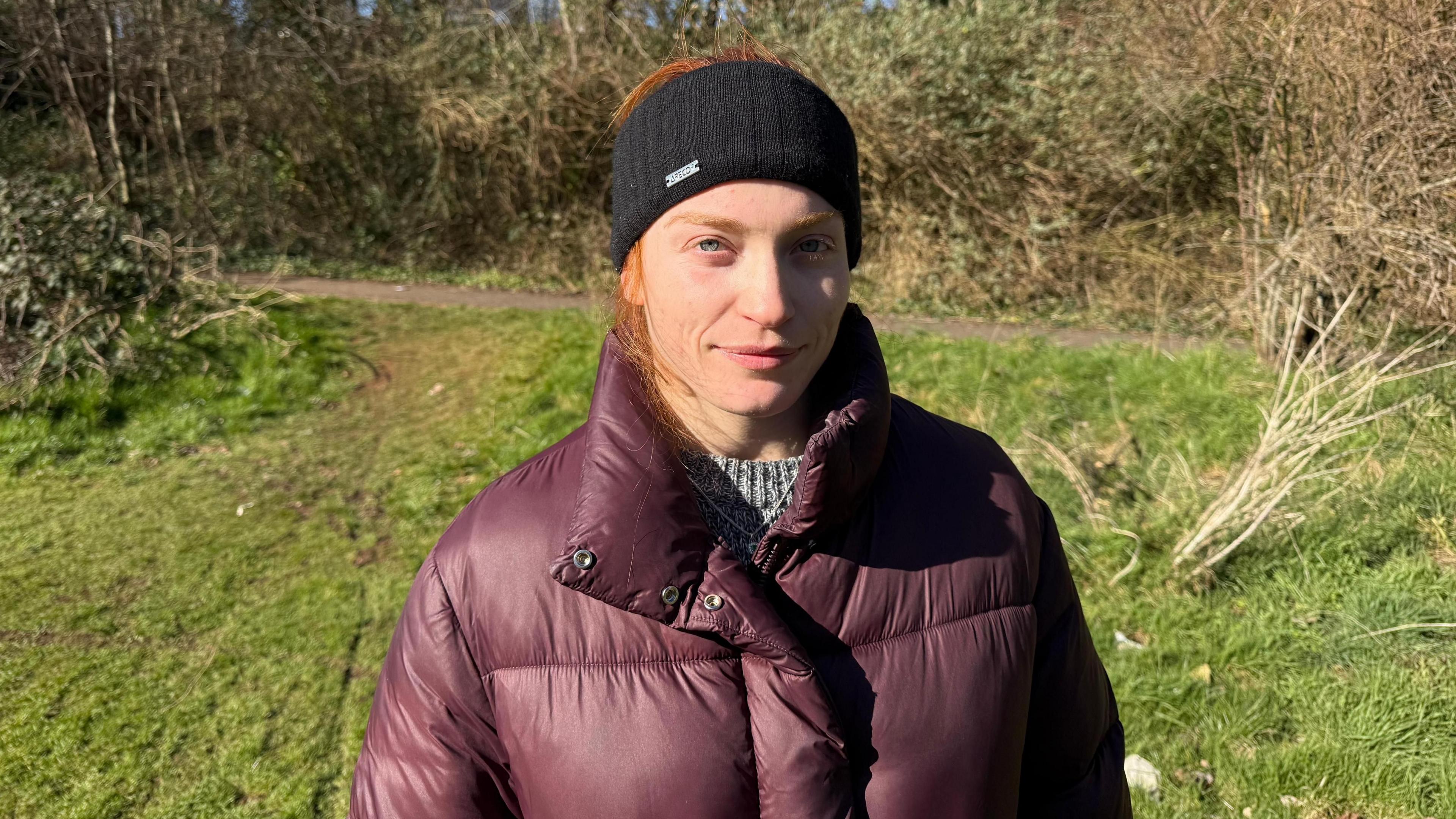 Lubjana has bright orange hair, most of which is tucked behind a thick black headband, and is wearing a thick purple puffer coat and grey jumper. She is smiling slightly at the camera, in a park. 