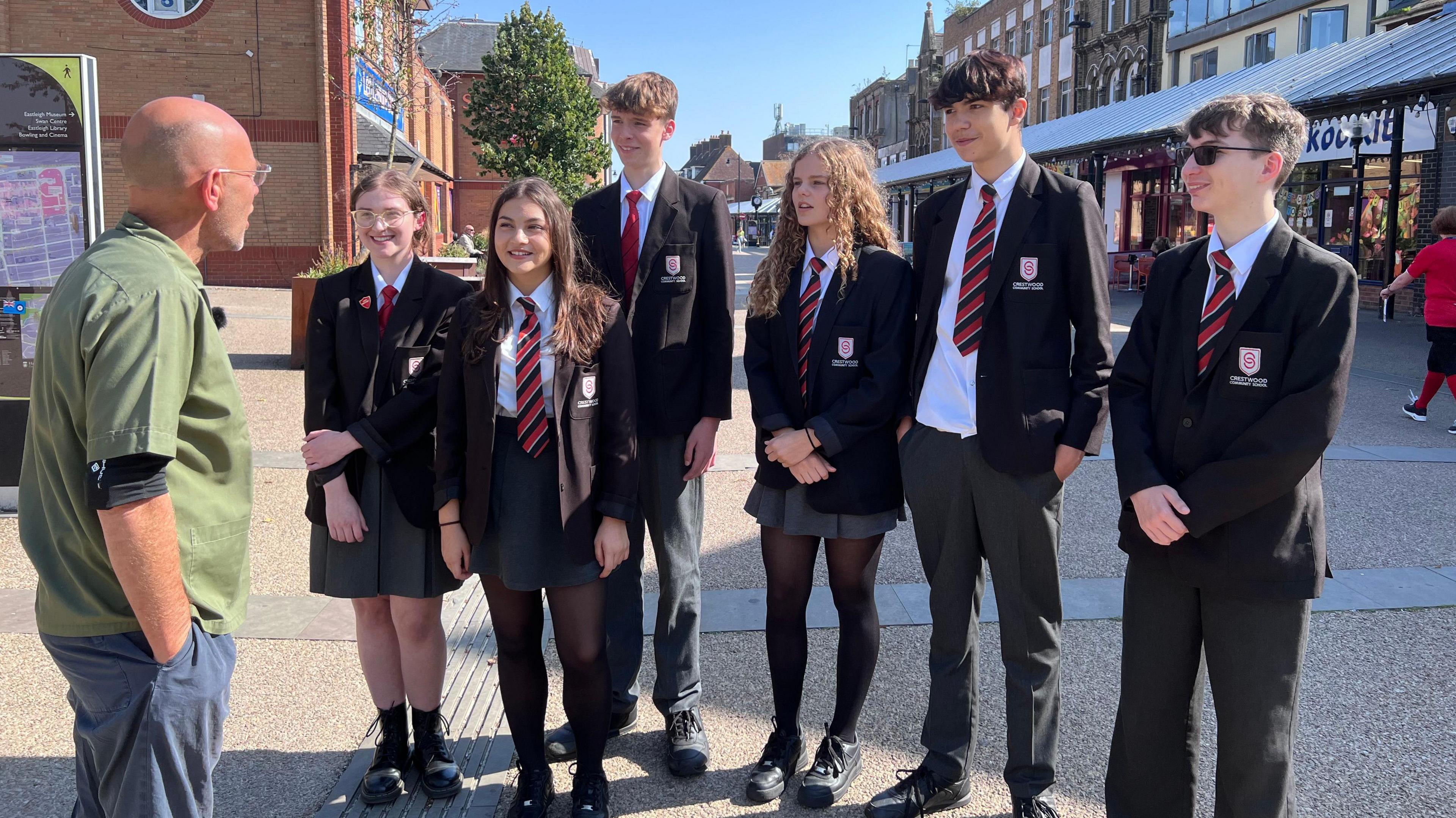 The designer Wayne Hemingway, wearing a green top, talking to six students - three boys and three girls in school uniform, outside some shops in Eastleigh