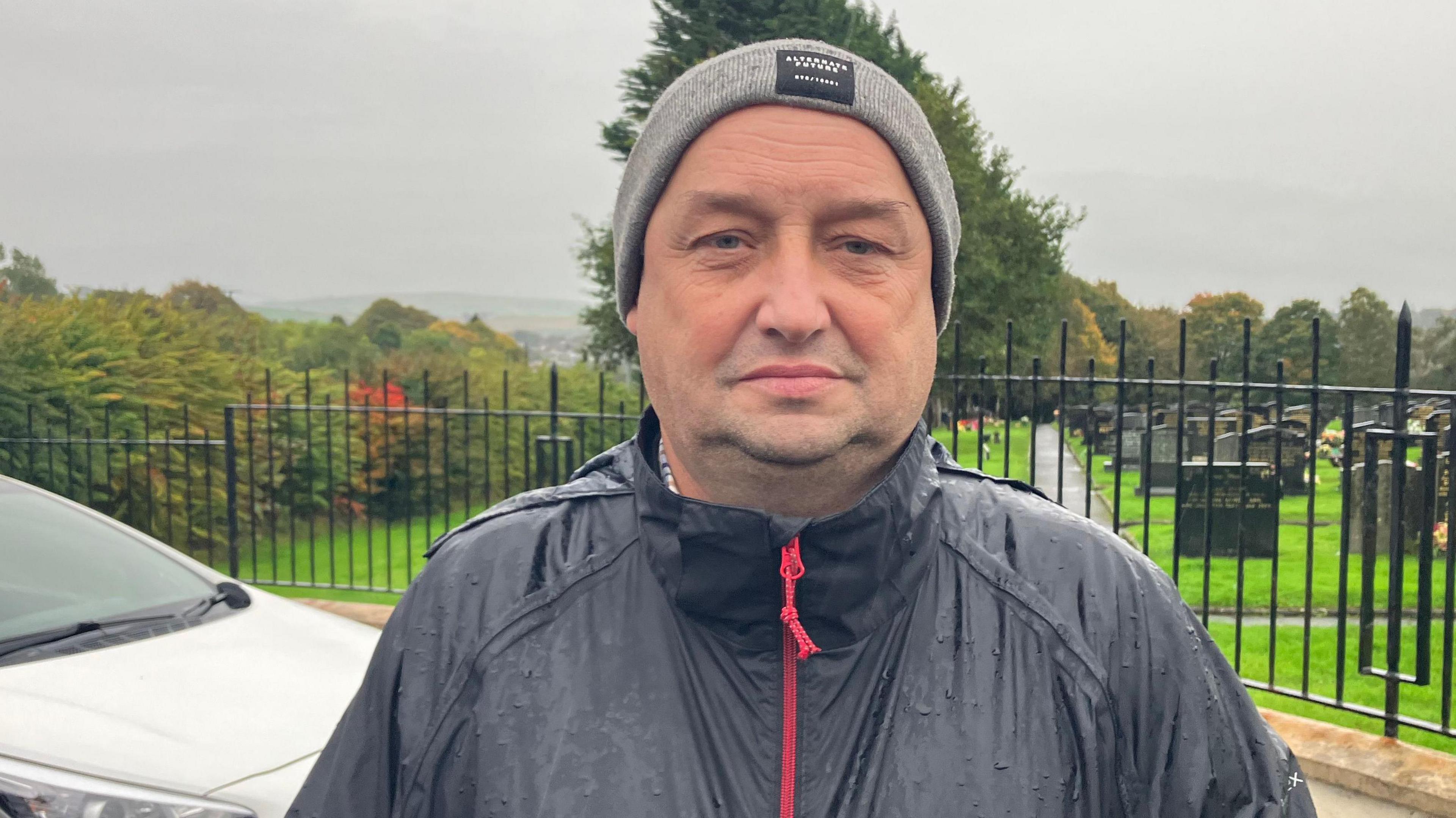 Declan Norris standing outside a cemetery, there are headstones behind him. He is wearing a black coat and a grey beanie hat. It's raining and the sky is grey due to Storm Amy.
