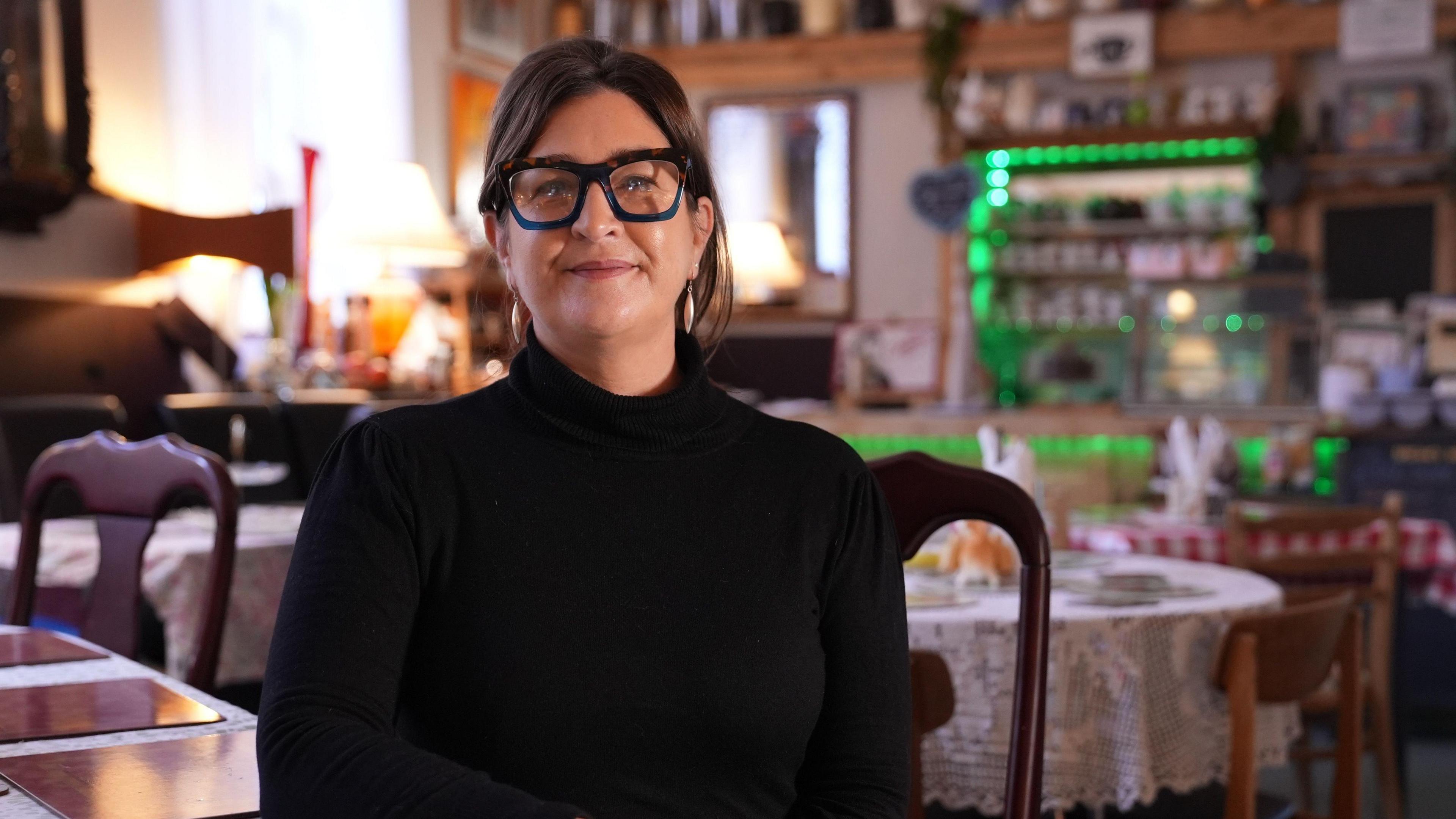 A woman with brown hair and glasses, wearing a black turtle neck jumper and sitting in a community cafe