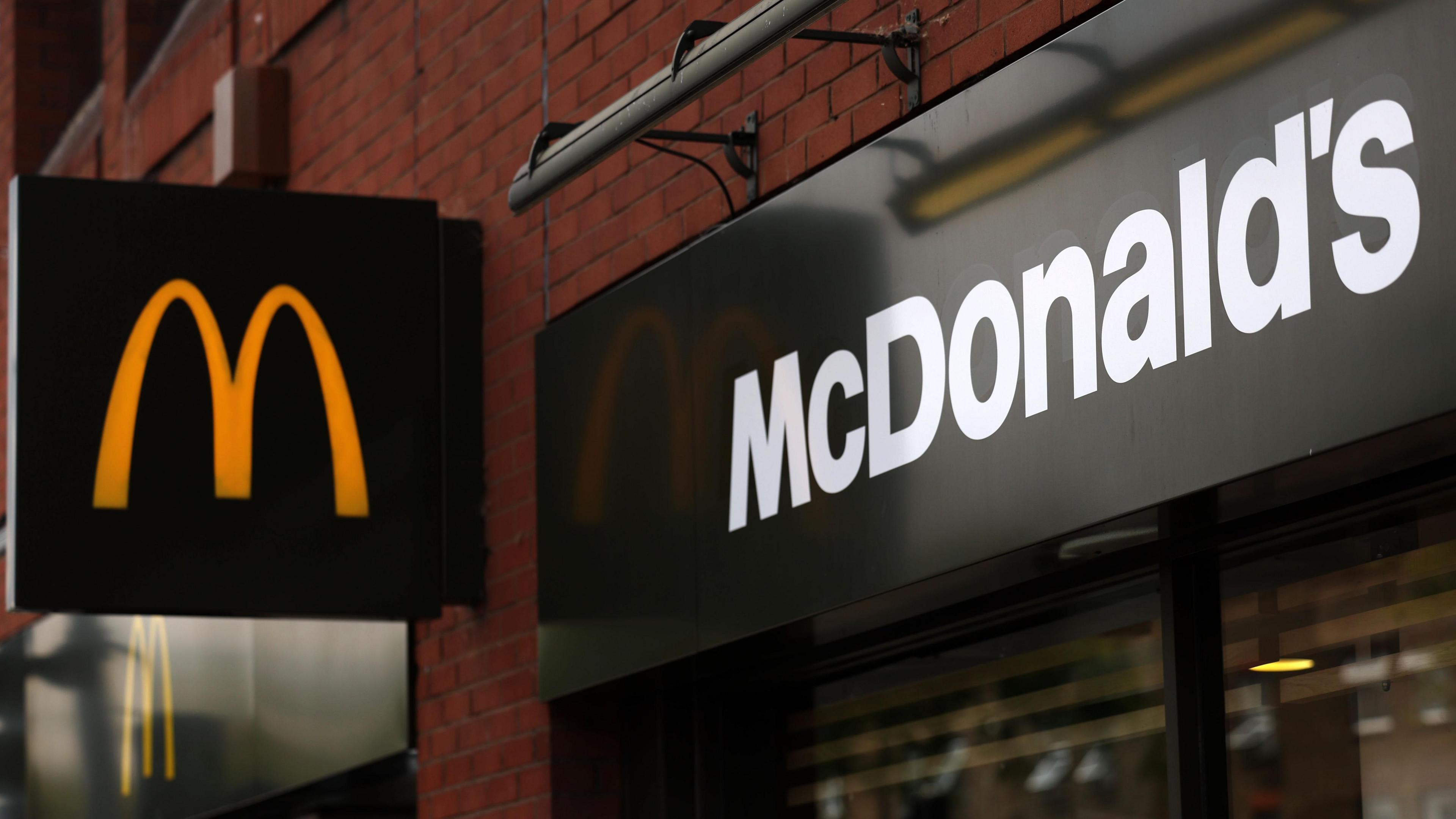 A exterior shot of a McDonald's restaurant with a black sign displaying a yellow "M" on it, with the full "McDonald's" title written above the entrance. 