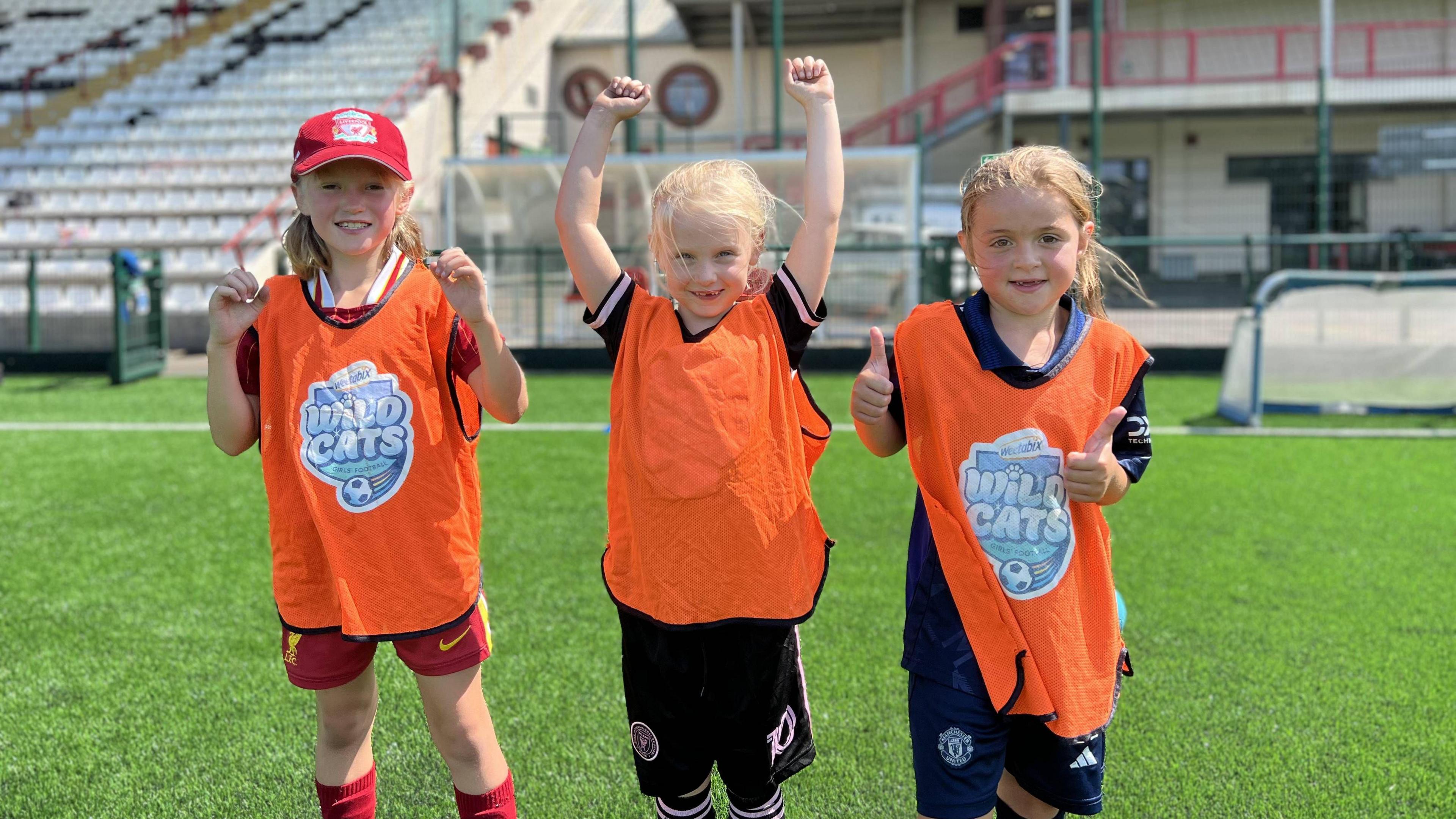 Three happy young girls in orange Wild Cats bibs cheer on a football pitch.