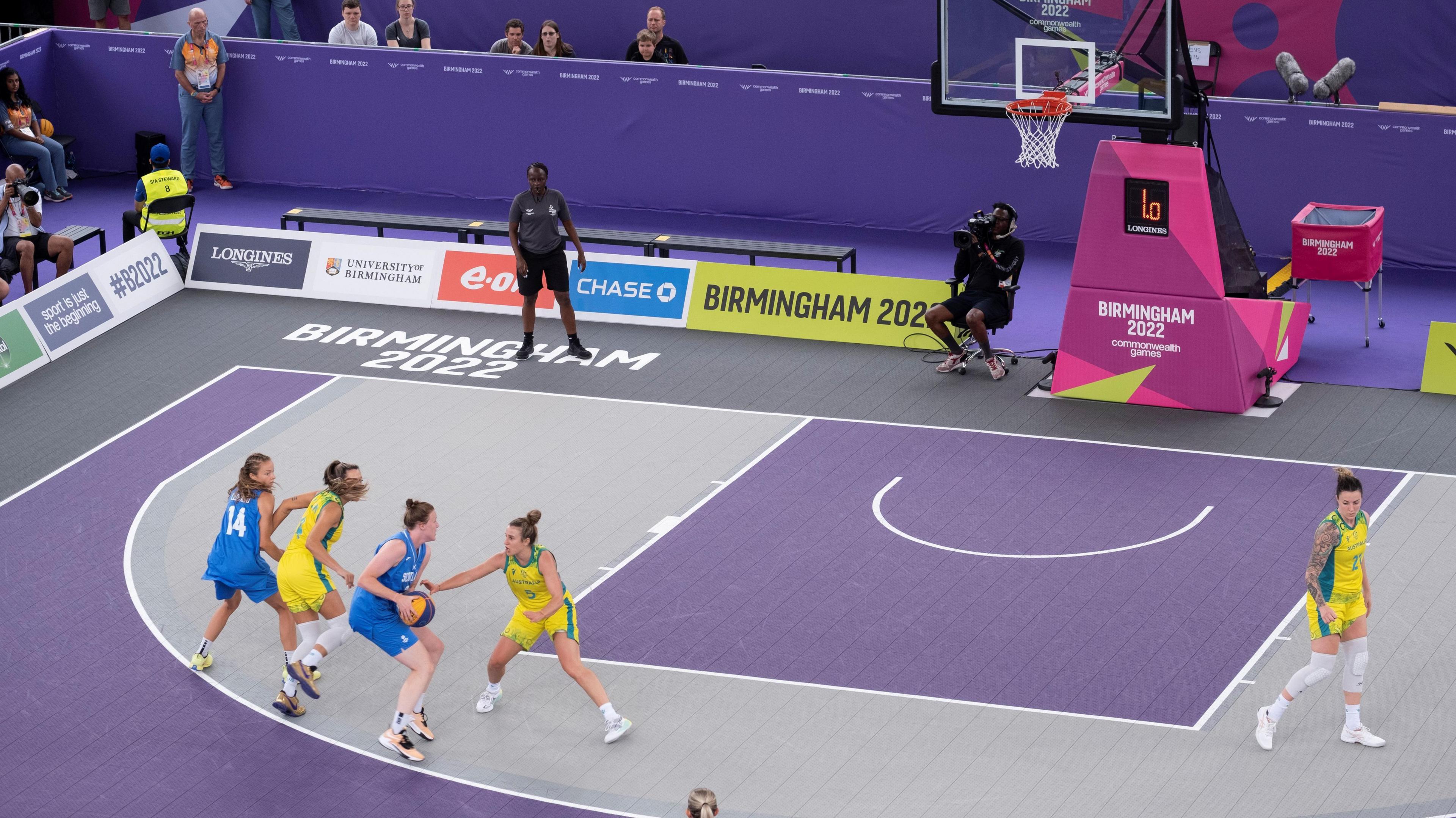 A basketball match taking place on a grey and purple court. The two female teams are wearing blue and yellow, with the blue team in possession of the ball.