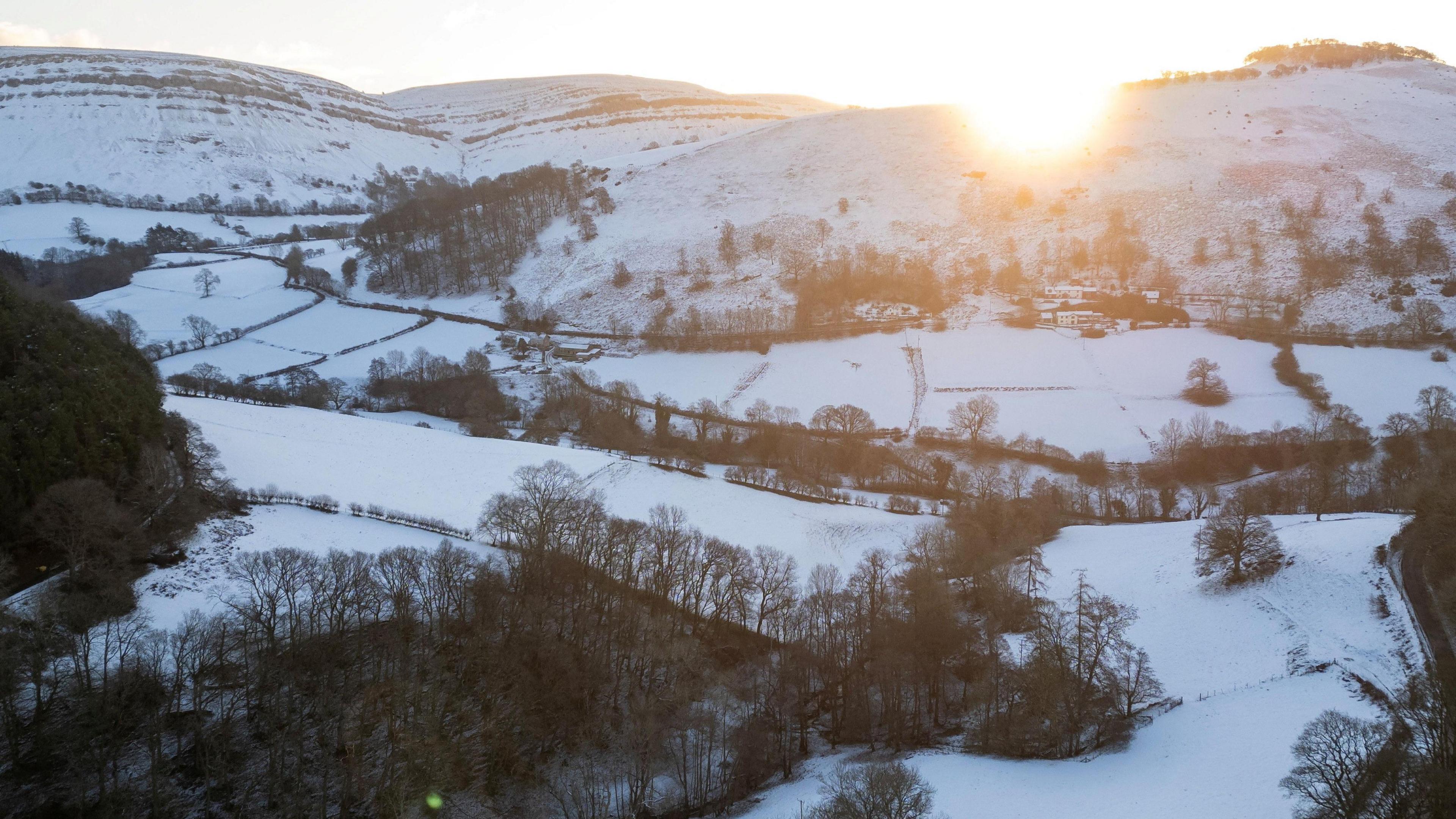 Hills and fields near Llangollen are covered in snow. The sun is shining over the top of the hill.