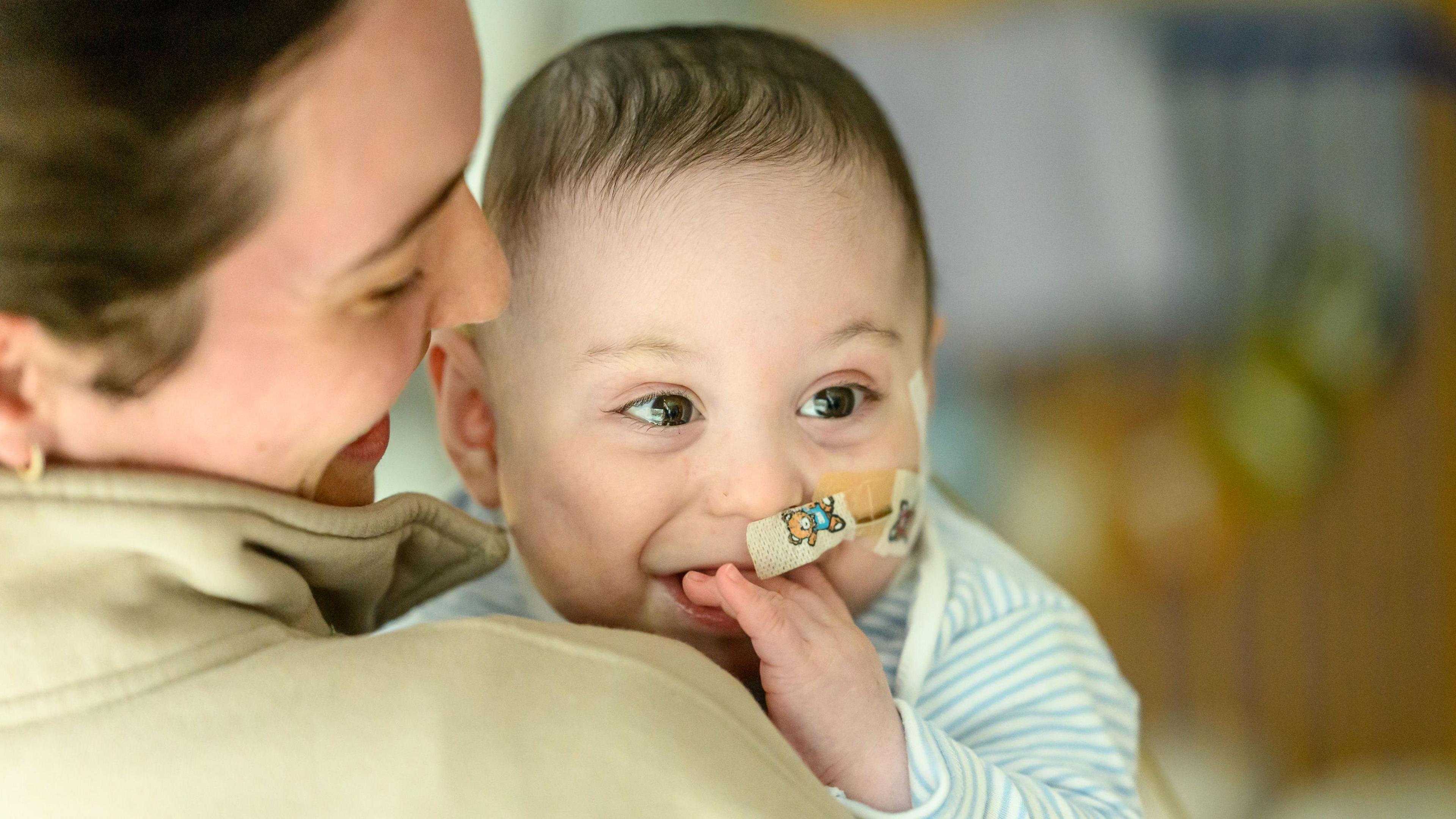 A young baby with dark hair looks past the camera, smiling. He has a feeding tube inserted into his nose and is chewing on of his hands.
A dark haired woman is holding him and she is pictured from behind.