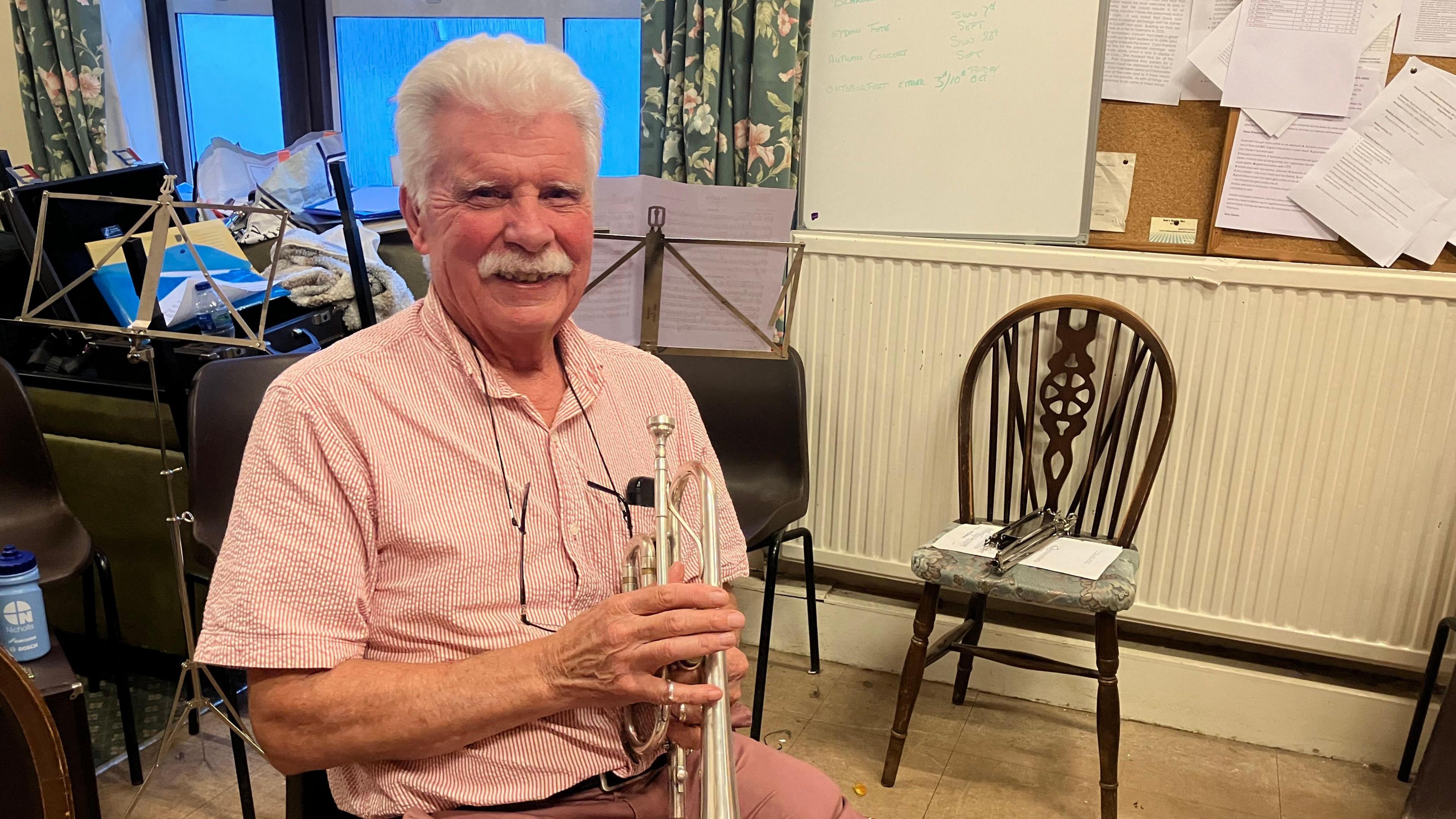 A man in a pink short sleeve shirt and pink trousers holds a cornet while seated smiling at the camera. 