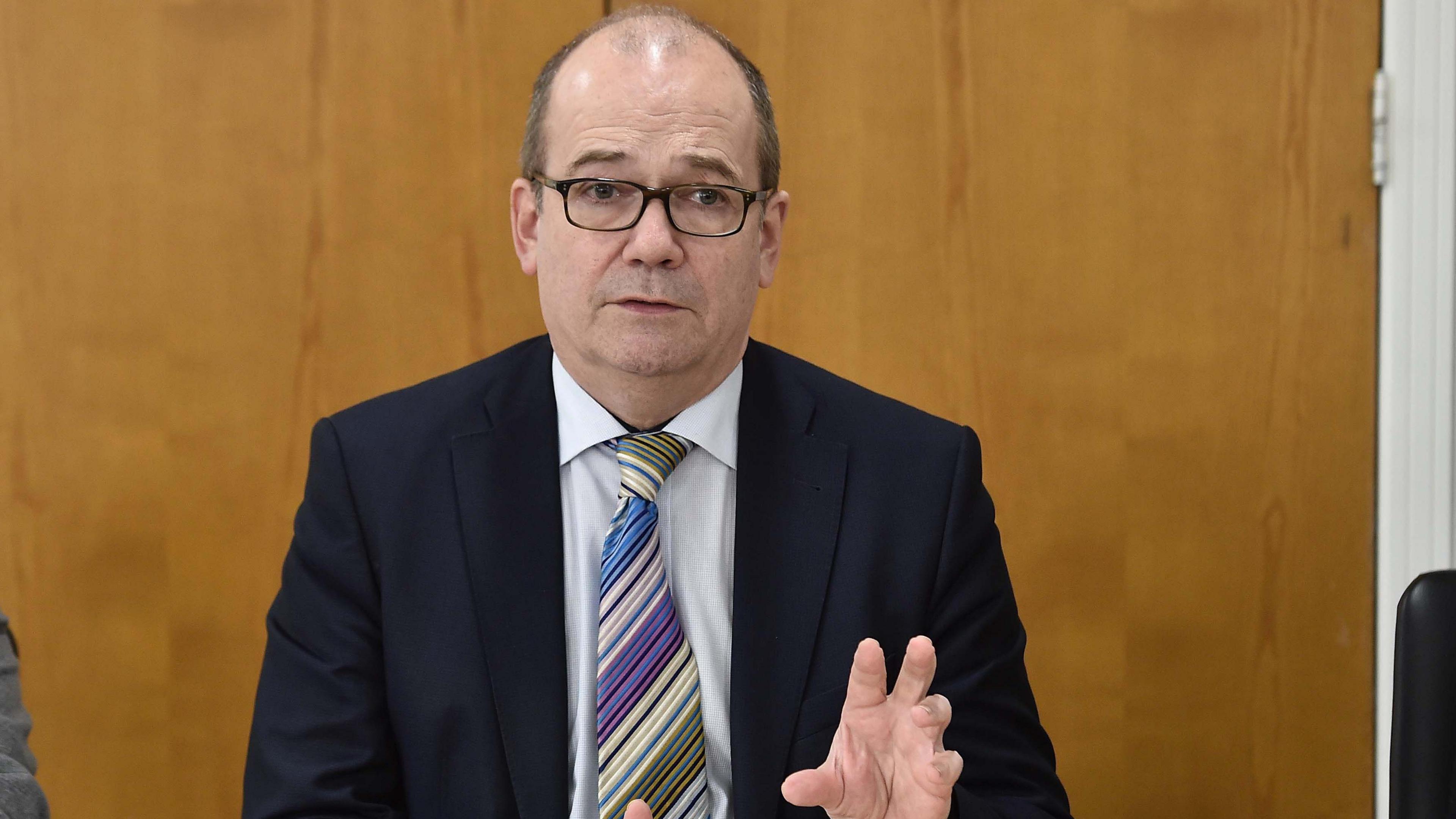 A man is sitting at a table. He wears glasses and has a black suit on and a stripped tie.